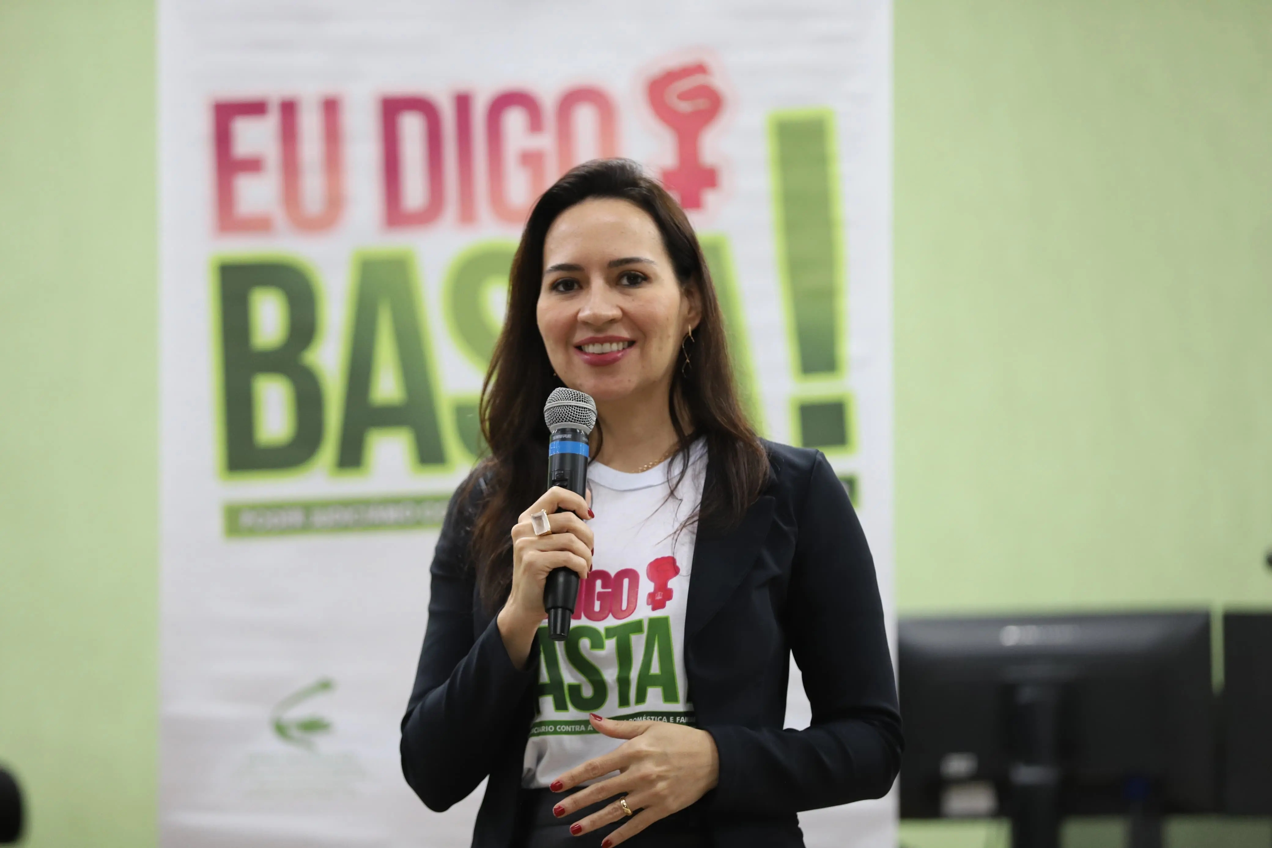 Mulher segura microfone e sorri, com camiseta da campanha “Eu Digo Basta!” e blazer preto. Ao fundo, banner da campanha com letras verdes e rosa sobre fundo claro.