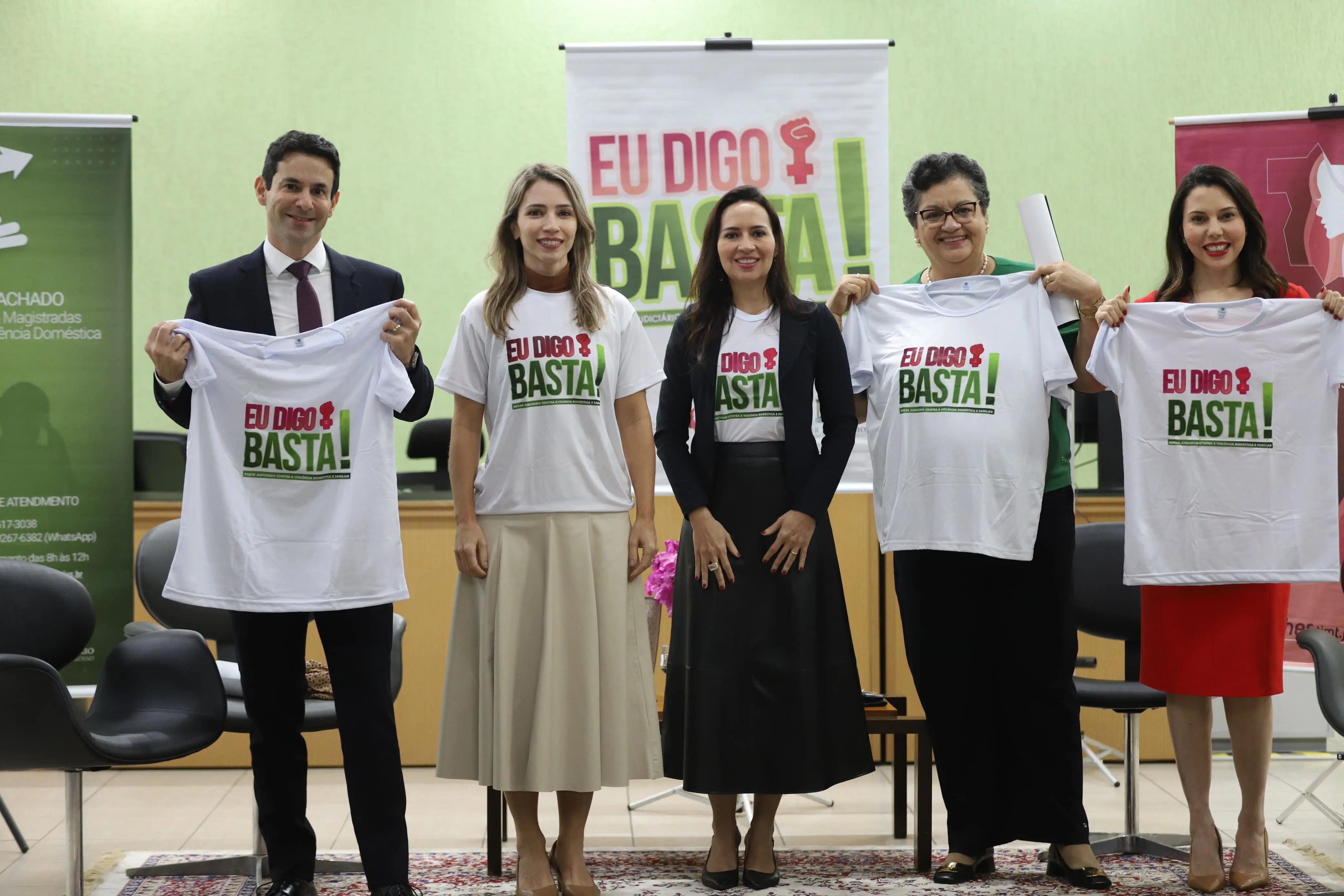 Cinco pessoas posam lado a lado, sorrindo, com camisetas “Eu Digo Basta!”. Ao fundo, banners da campanha e cenário de evento oficial com flores no centro do palco.