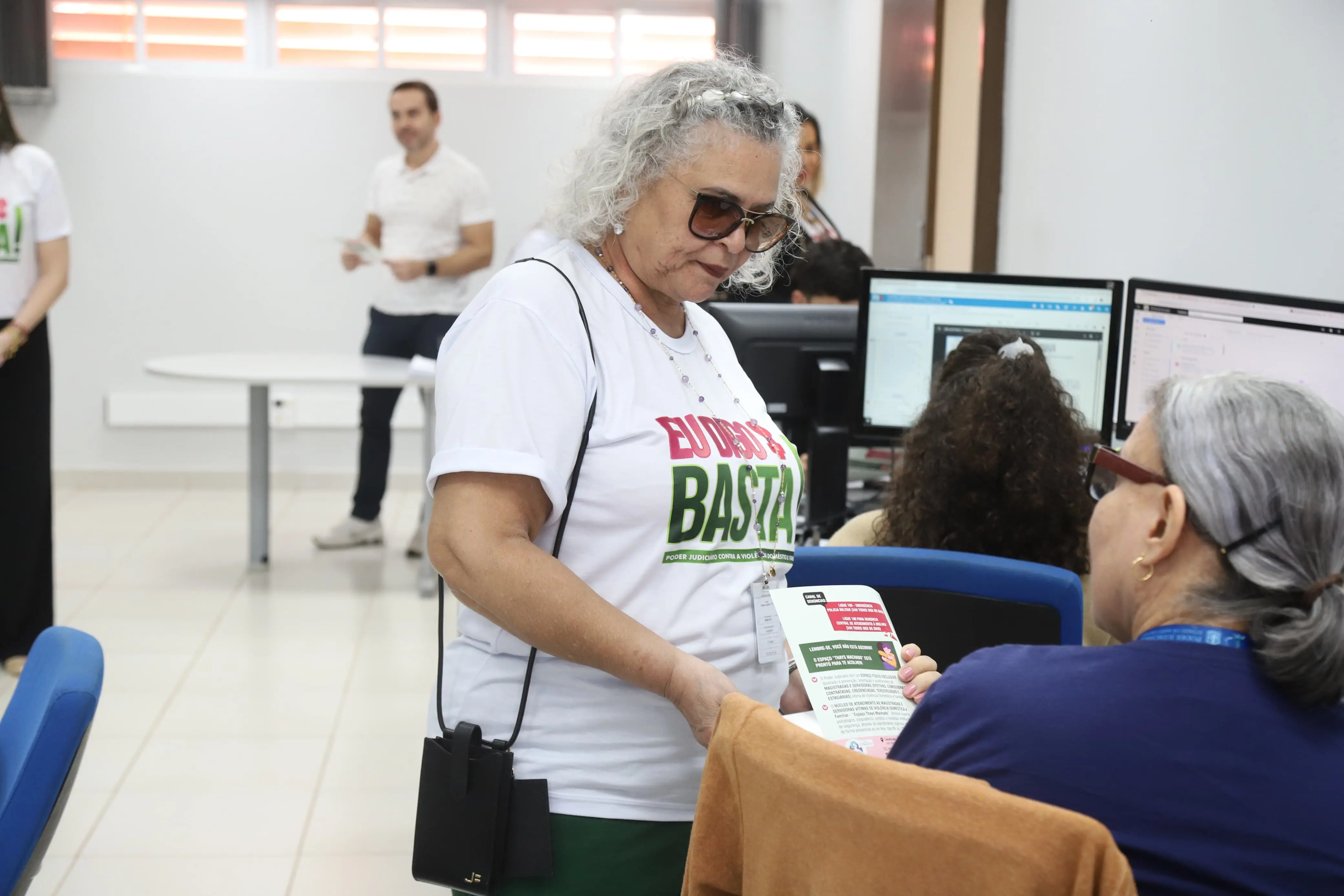 Uma mulher de cabelos grisalhos e óculos escuros, vestindo camiseta "Eu Digo Basta!", entrega um folheto a outra pessoa sentada em um escritório com computadores. A cena reflete interação e compartilhamento de informações, promovendo a inclusão e a conscientização.