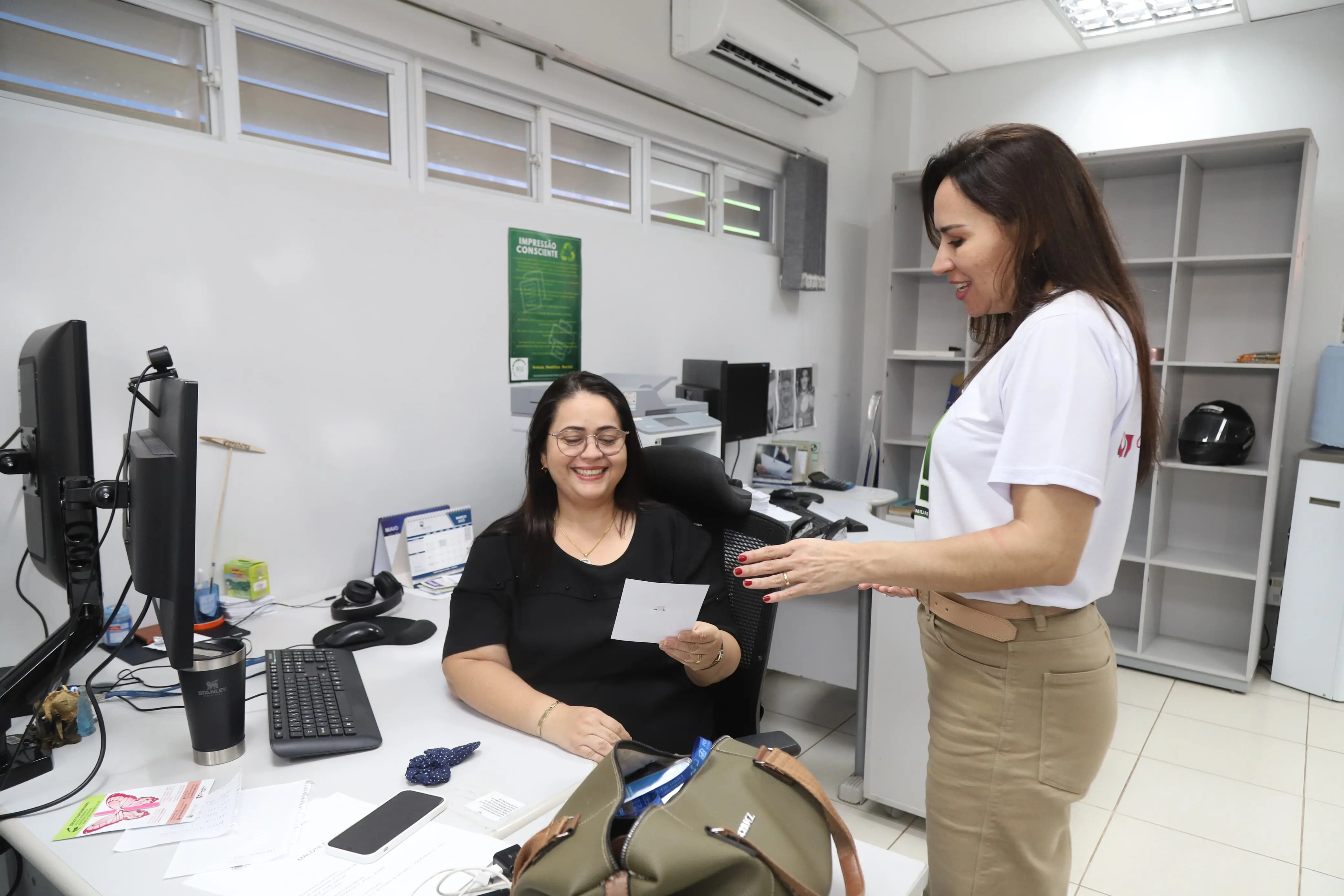 uma mulher sorridente sentada em sua mesa de escritório, usando óculos e blusa preta, segura um pequeno papel. Outra mulher, de cabelos longos e camiseta branca, está de pé ao lado da mesa, com as mãos abertas, em atitude de diálogo. A cena mostra interação e troca de informações em um ambiente de t
