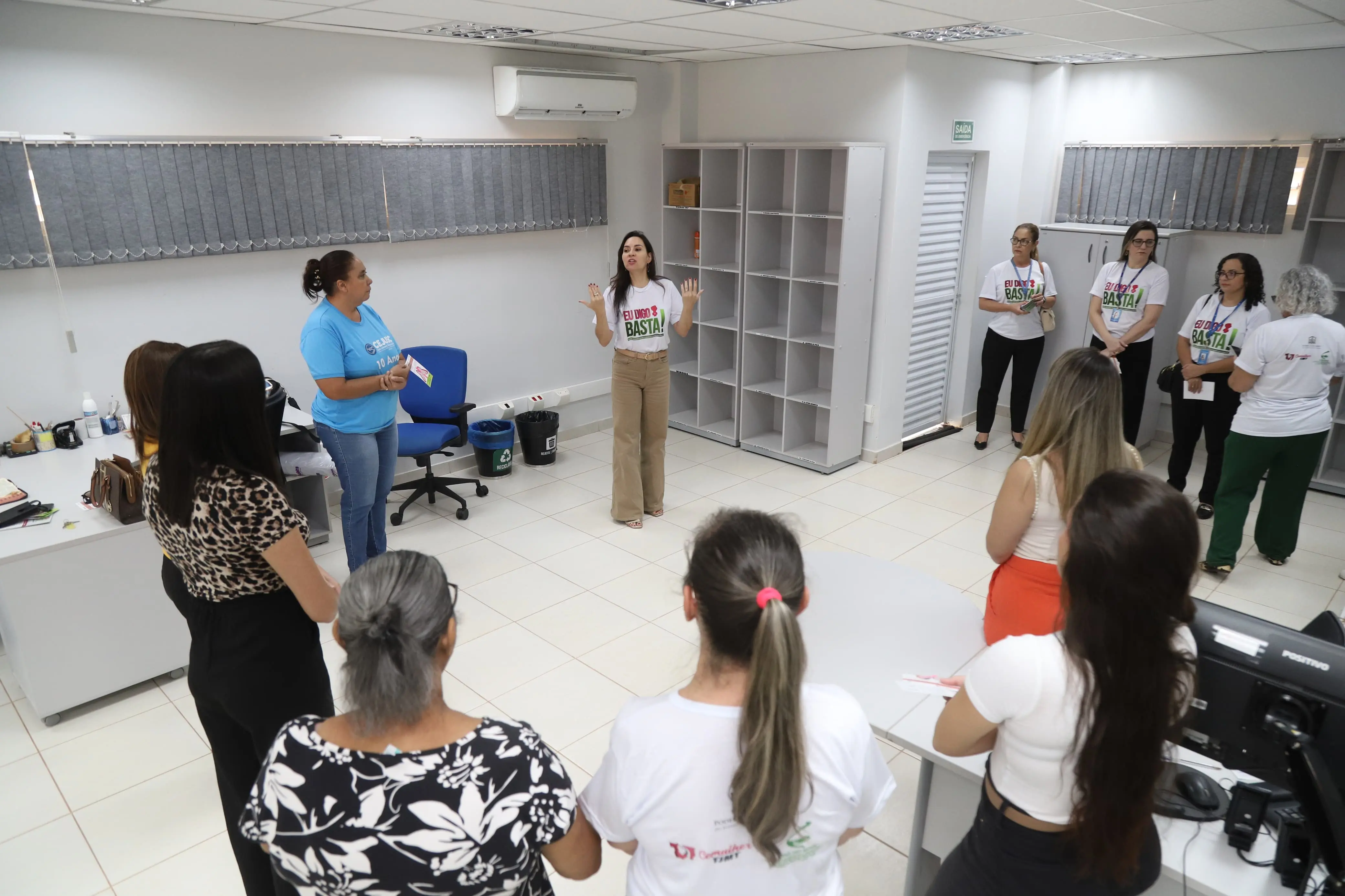 Uma mulher de calça bege e camiseta "Eu Digo Basta!" está no centro de uma sala, gesticulando para um grupo de mulheres que a ouvem atentas. O ambiente de escritório indica uma reunião ou palestra