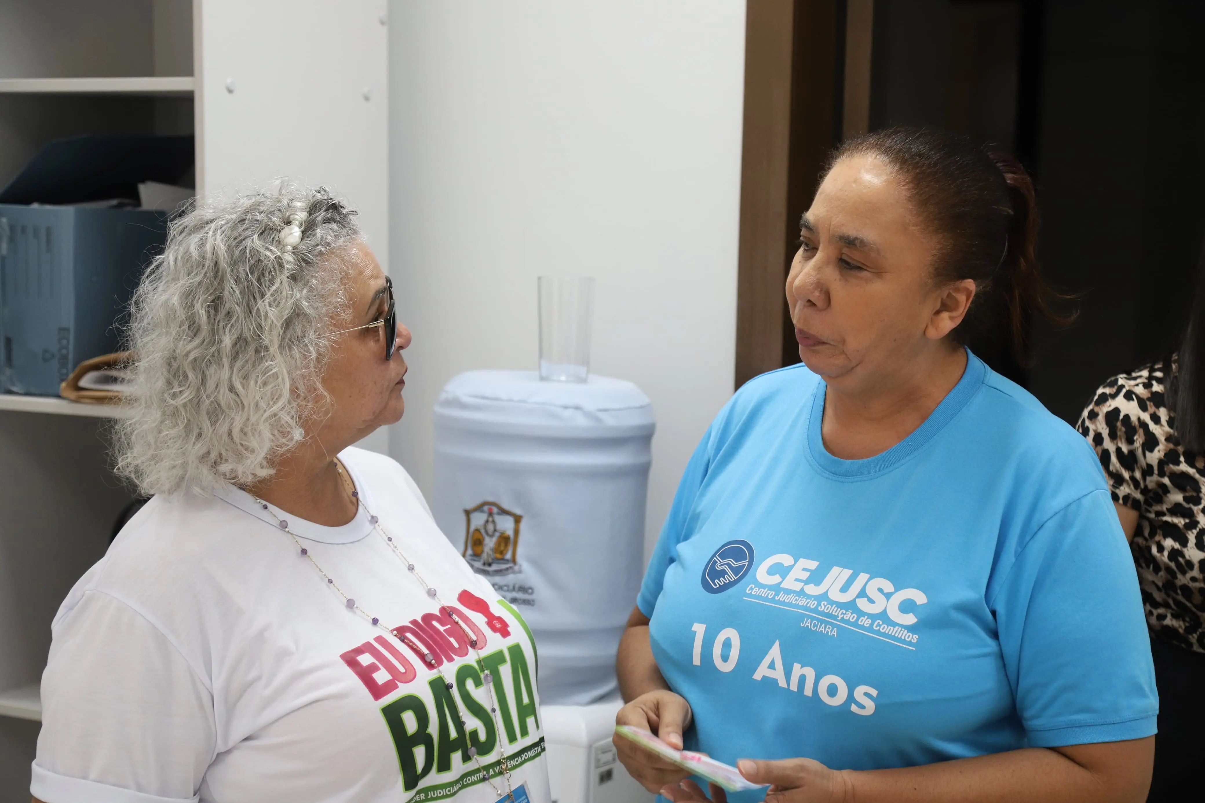 Duas mulheres conversam em um ambiente de escritório. Uma, de cabelos grisalhos e camiseta "Eu Digo Basta!", olha atenta para a outra, de camiseta azul "CEJUSC 10 Anos", que segura algo nas mãos. 