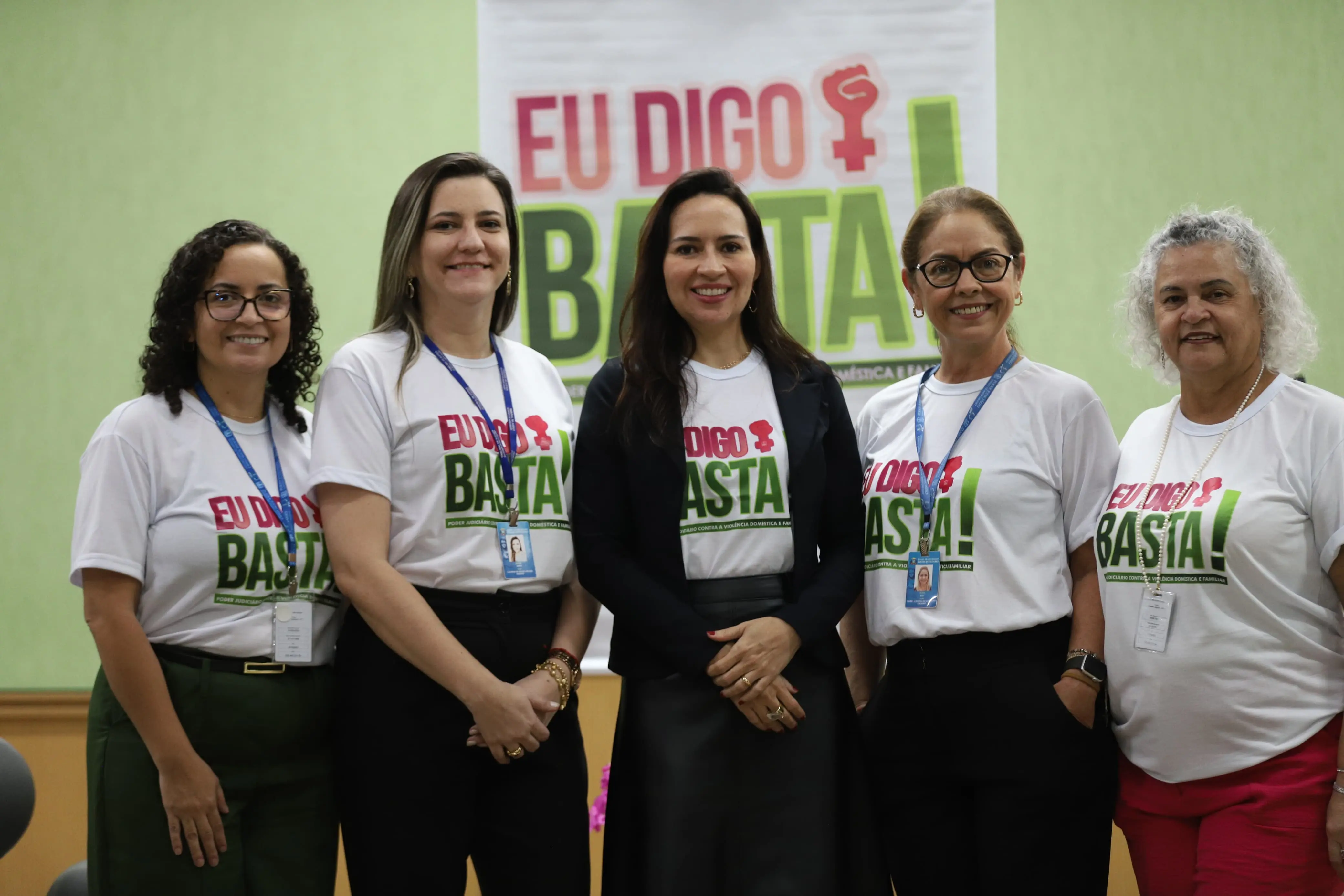 Cinco mulheres posam lado a lado com sorrisos. Usam camisetas “Eu Digo Basta!” e crachás. Ao fundo, banner da campanha. Ambiente claro e institucional.