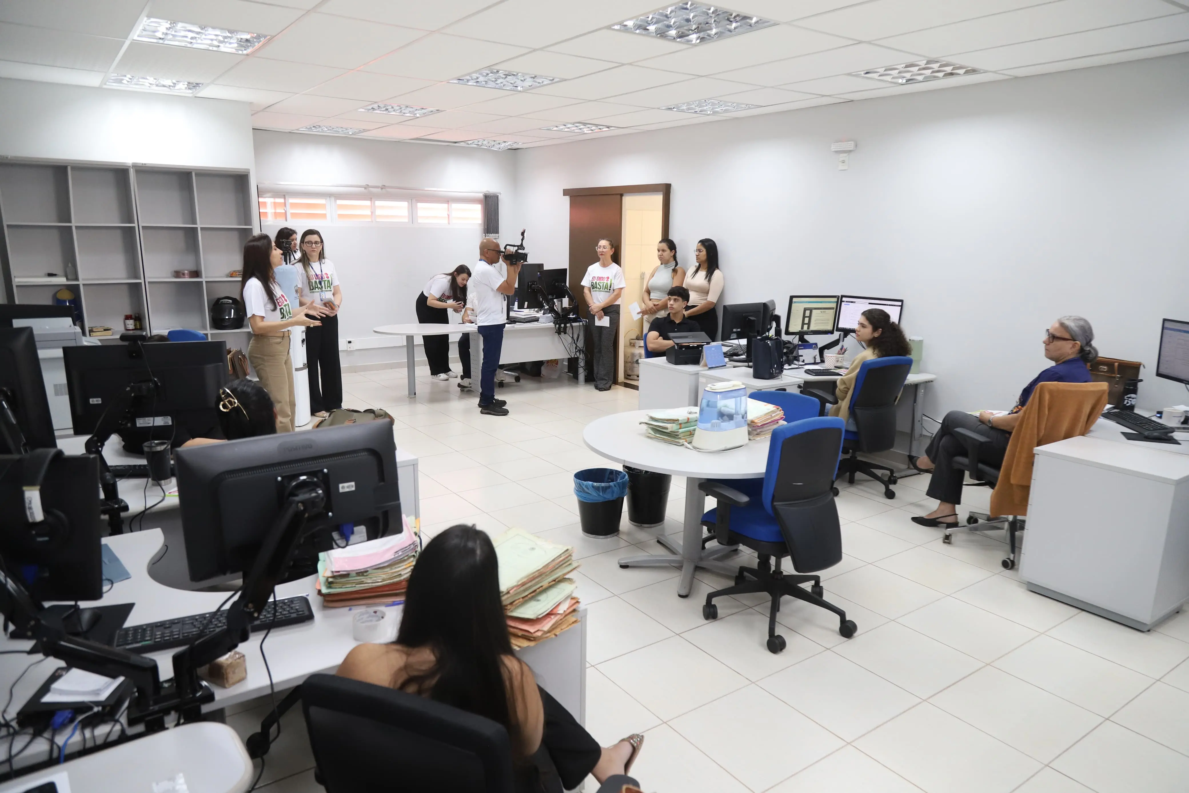 várias pessoas trabalham em um escritório moderno, com mesas, cadeiras e computadores. Um grupo, incluindo um homem com câmera, parece registrar um evento da campanha "Eu Digo Basta!", com alguns usando as camisetas da iniciativa.