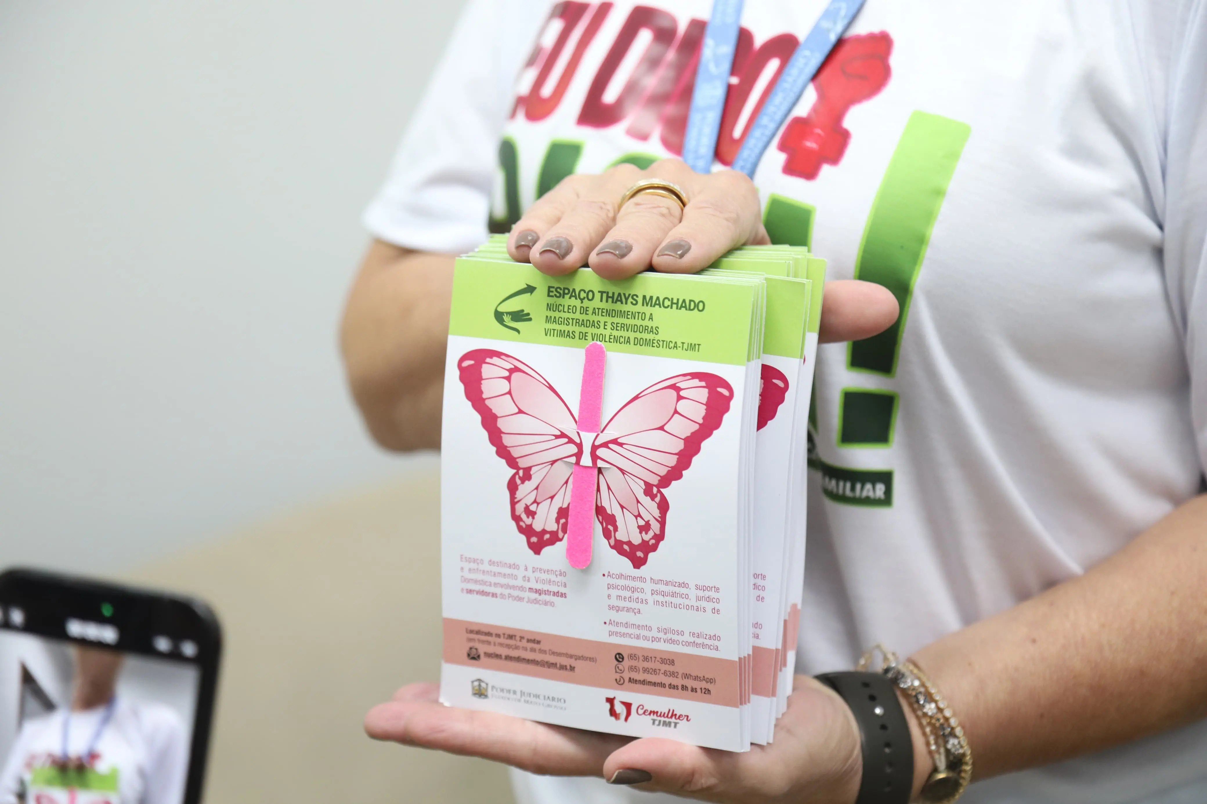 Uma pessoa segura folhetos da campanha "Espaço Thays Machado", com uma borboleta rosa e verde. Os folhetos informam sobre apoio a vítimas de violência doméstica, como juízas e servidoras, promovendo a conscientização e o acesso a serviços. A imagem sugere a distribuição de materiais informativos.