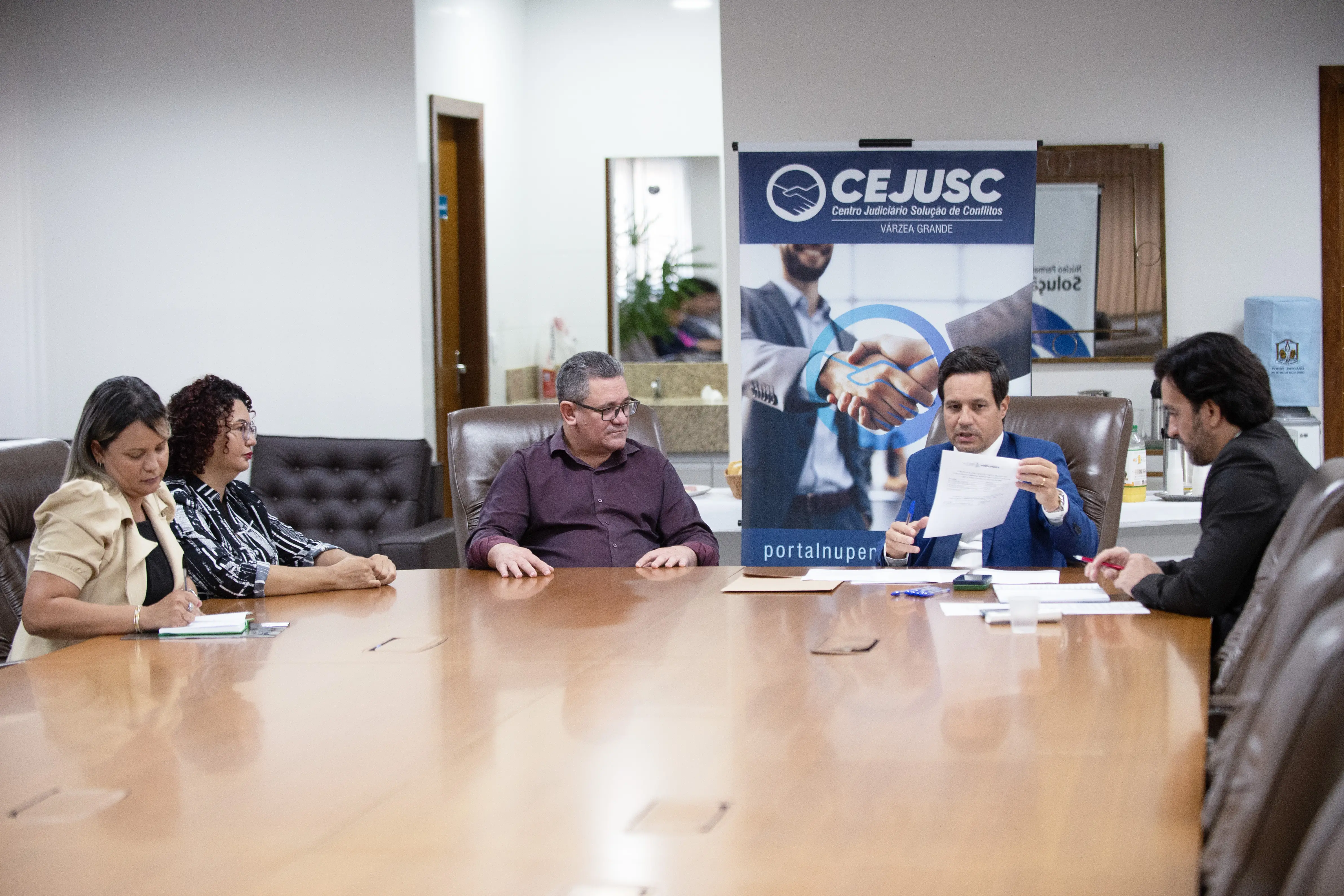 Foto em plano aberto que mostra duas mulheres e três homens (servidoras e secretário da Secretaria de Educação de Várzea Grande, juiz e assessor) sentados e conversando em volta de uma mesa na sala de reuniões do Fórum de Várzea Grande. 