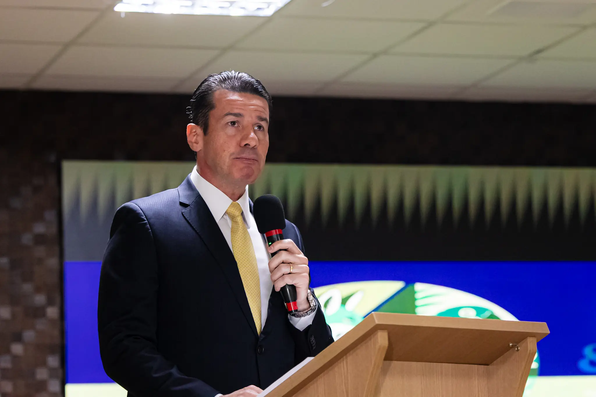 Homem de terno escuro, gravata amarela e cabelos pretos penteados para trás discursa em um púlpito de madeira enquanto segura um microfone preto. Ao fundo, um painel azul e verde.