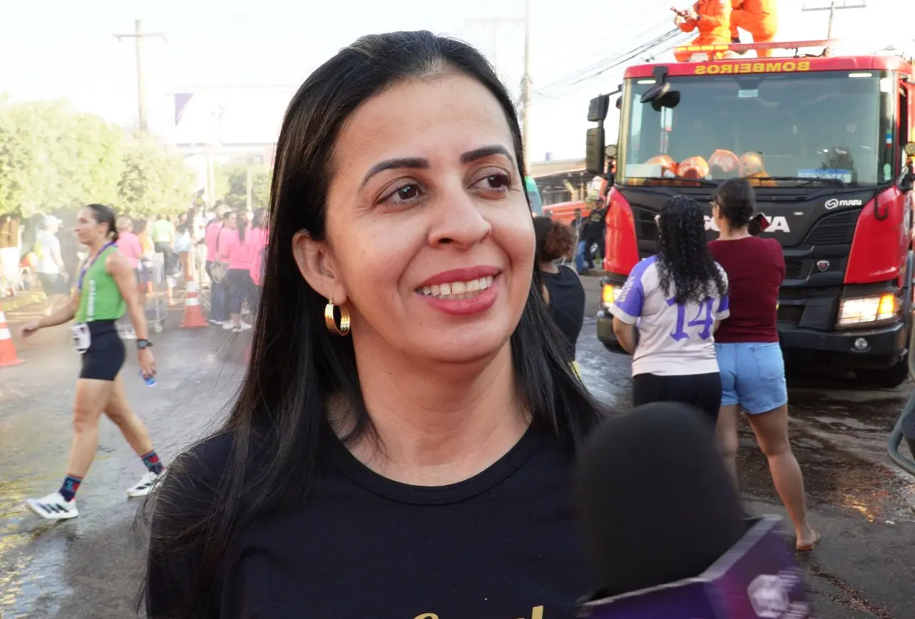 Uma mulher sorri durante entrevista concedida a microfone, em via pública tomada por participantes de prova esportiva. Ao fundo, viatura dos bombeiros e público acompanham o evento.