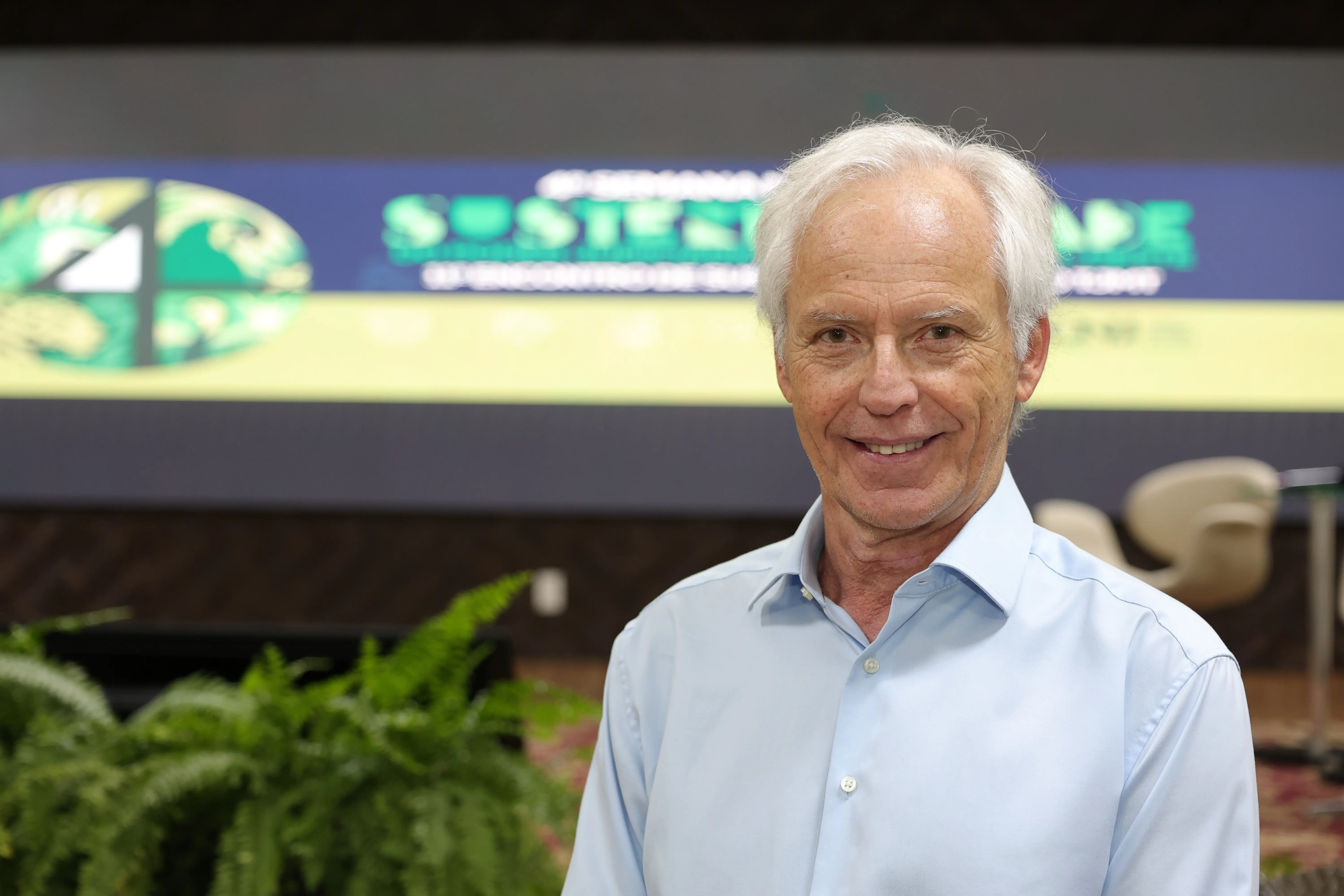 Um homem de cabelos grisalhos e camisa azul clara sorri para a câmera. O mesmo telão com o logo verde e a palavra "SUSTENTABILIDADE" está ao fundo, desfocado.