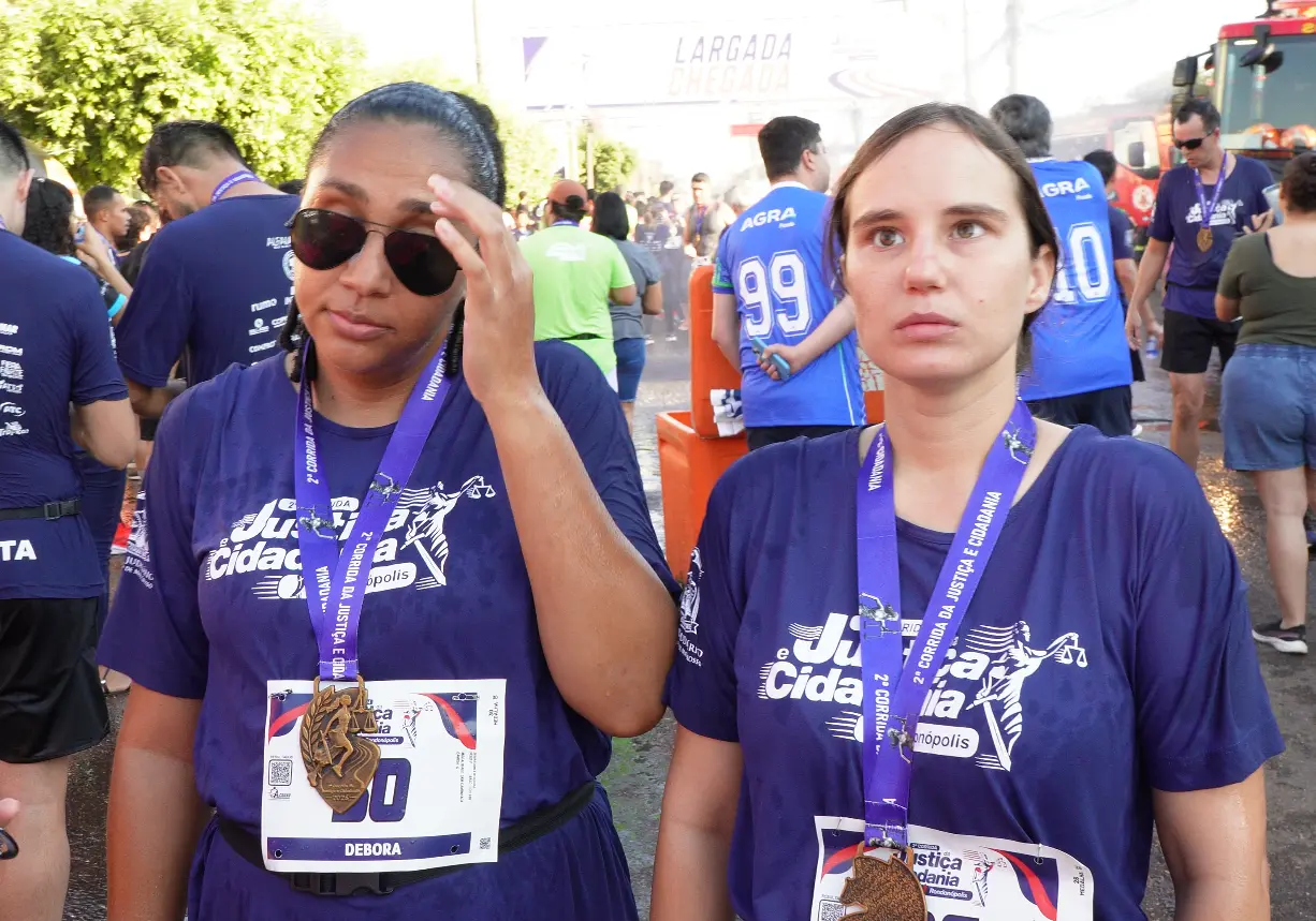Duas mulheres posam lado a lado exibindo medalhas de participação em prova esportiva, com números de peito identificados. Ao fundo, participantes se aglomeram próximo ao arco de chegada.
