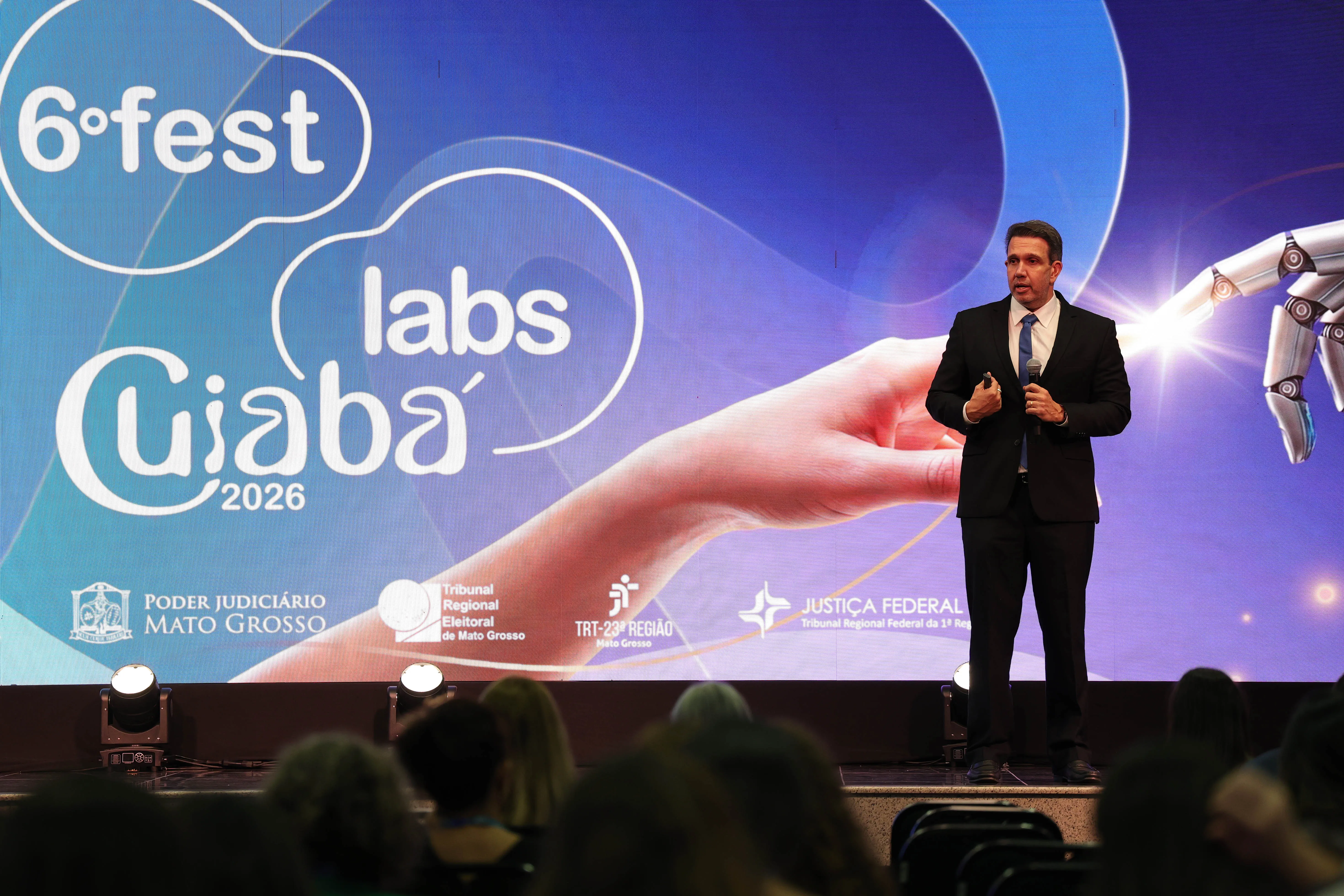 Homem de terno preto e gravata azul gesticula no palco diante de um telão azul com a inscrição "6º fest labs Cuiabá 2026" e grafismos futuristas. Na plateia, em primeiro plano, veem-se cabeças de pessoas assistindo.