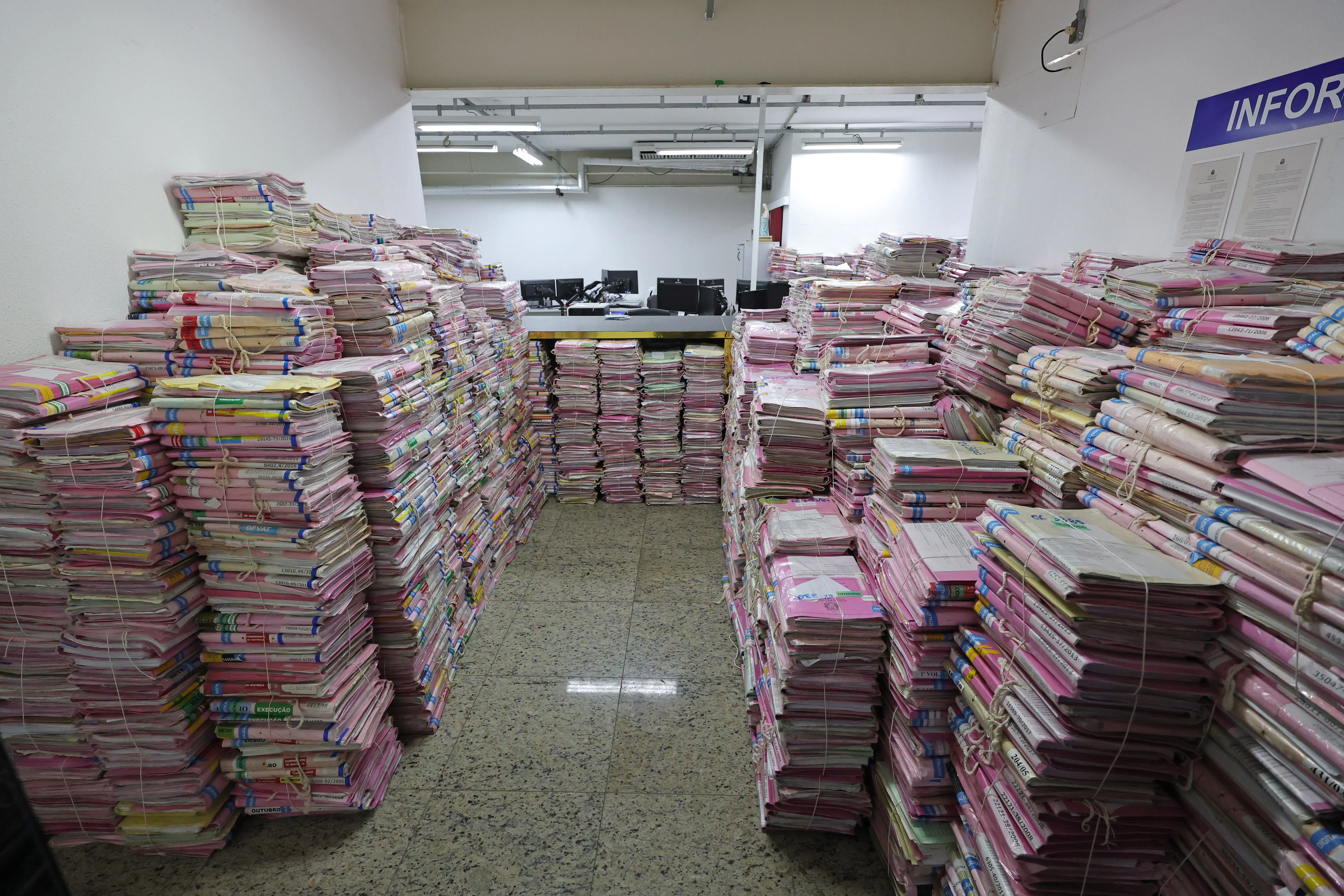 Corredor estreito formado por altas e numerosas pilhas de processos e pastas amarrados, a maioria em tons de rosa, que se estendem pelas laterais e ao fundo. Ao longe, vê-se uma divisória com computadores. No alto, à direita, há uma placa onde se lê parcialmente "INFOR".