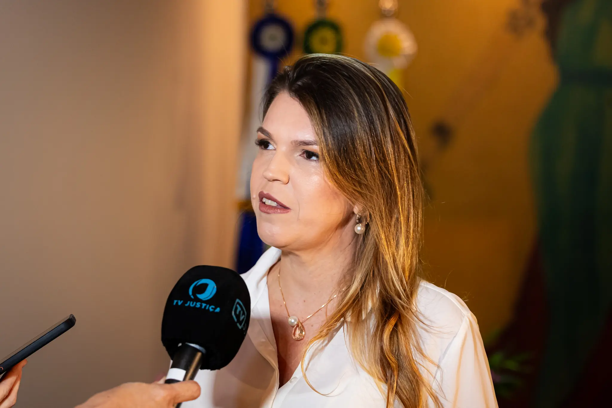 Mulher de pele clara e cabelos castanhos claros na altura dos ombros concede entrevista. Usa camisa branca e colar com pendente de pérola. À sua frente, um microfone preto com a marca "TV JUSTIÇA" em azul e branco.