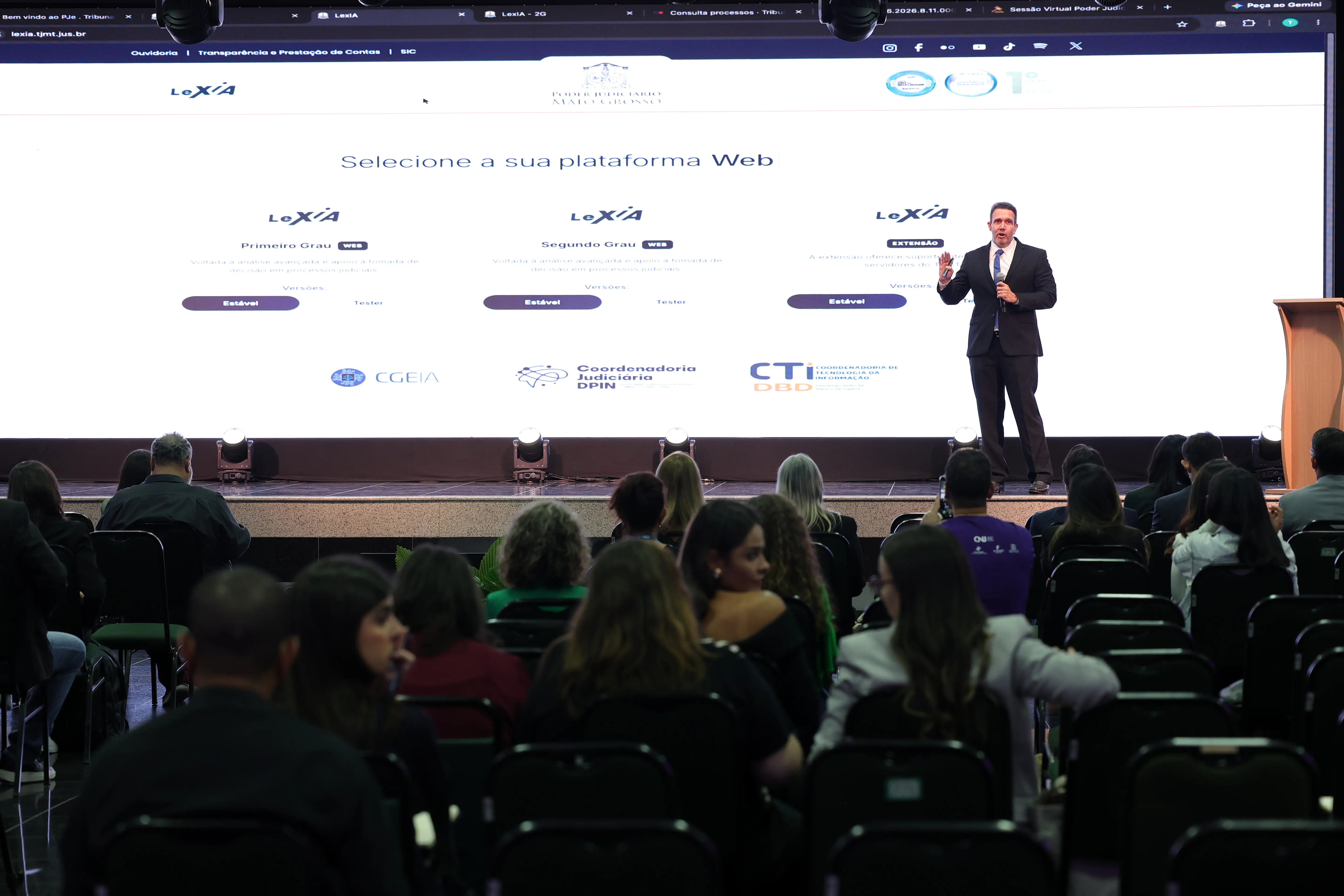 Visão geral de um auditório com plateia de costas. Ao fundo, no palco, um homem de terno fala com microfone ao lado de um grande telão branco que exibe o site do Poder Judiciário de Mato Grosso e o sistema "LeXIA".