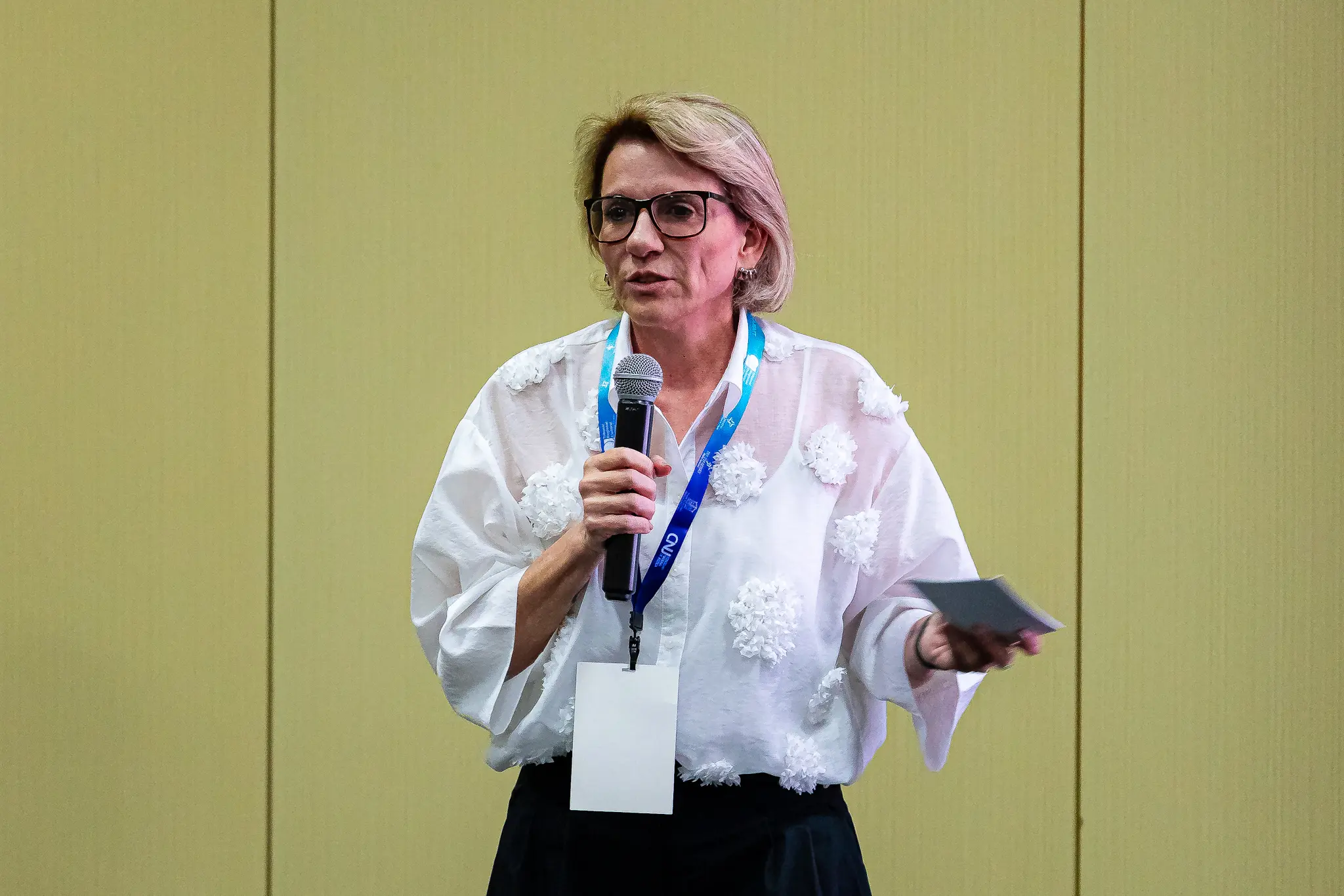 Daniella, mulher de cabelos claros e curtos, discursa ao microfone, de óculos, blusa branca florida e crachá do CNJ pendurado no pescoço, gesticula durante evento institucional.