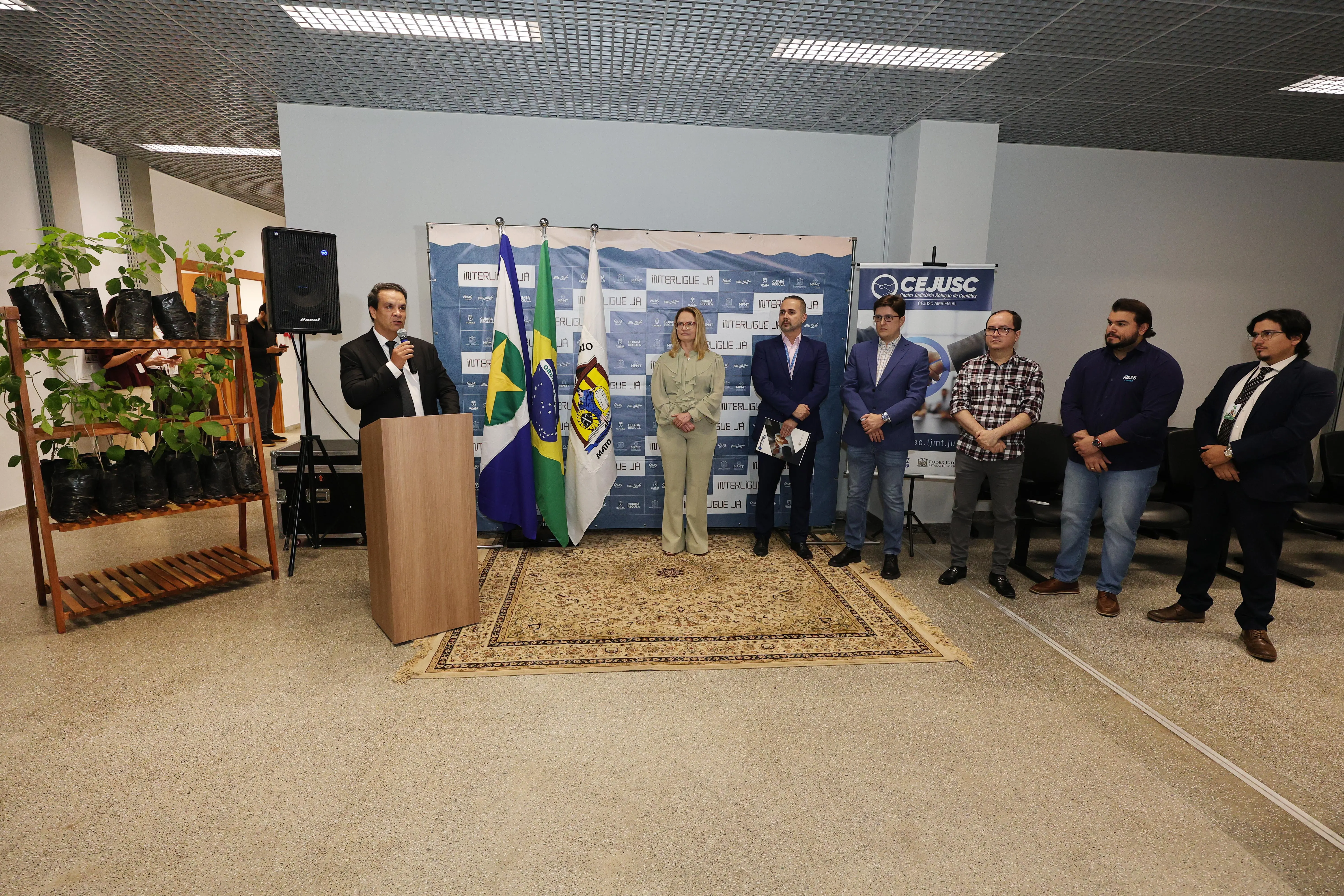 Homem discursa em púlpito ao lado de bandeiras e tapete. À sua direita, grupo de autoridades acompanha em pé. À esquerda, estante com mudas de plantas. Ao fundo, painel com a marca CEJUSC.