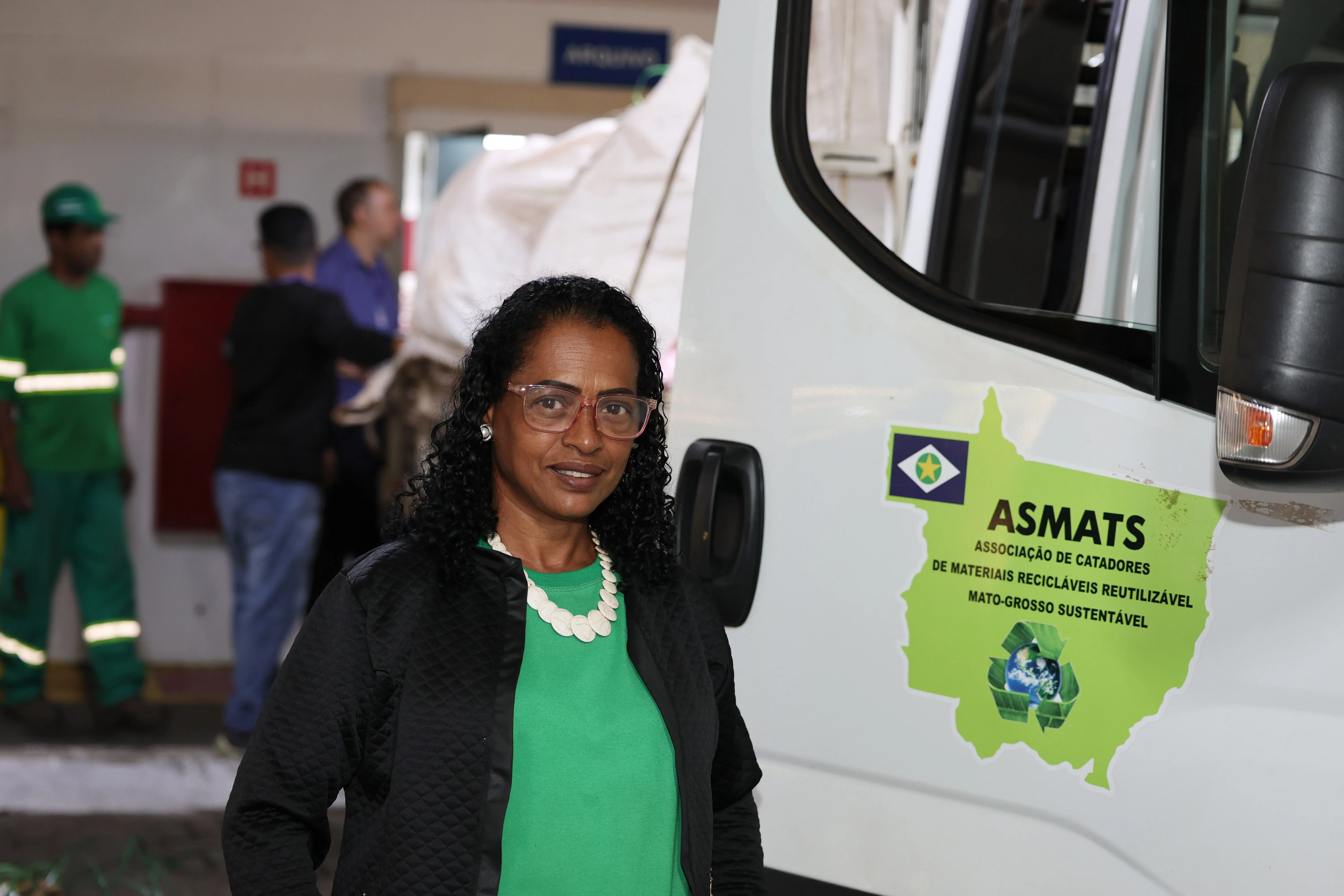 Mulher negra com óculos, colar claro, blusa verde e casaco preto sorri em primeiro plano. À direita, a lateral de um veículo branco com o logotipo "ASMATS" e texto sobre catadores. Ao fundo, em ambiente de galpão, homens trabalham perto de grandes fardos e outro veículo.