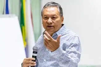 Homem de meia idade, cabelos grisalhos, vestindo camisa de botões azul claro com padronagem, está em pé e fala. Ele segura um microfone com a mão direita e gesticula com a esquerda.