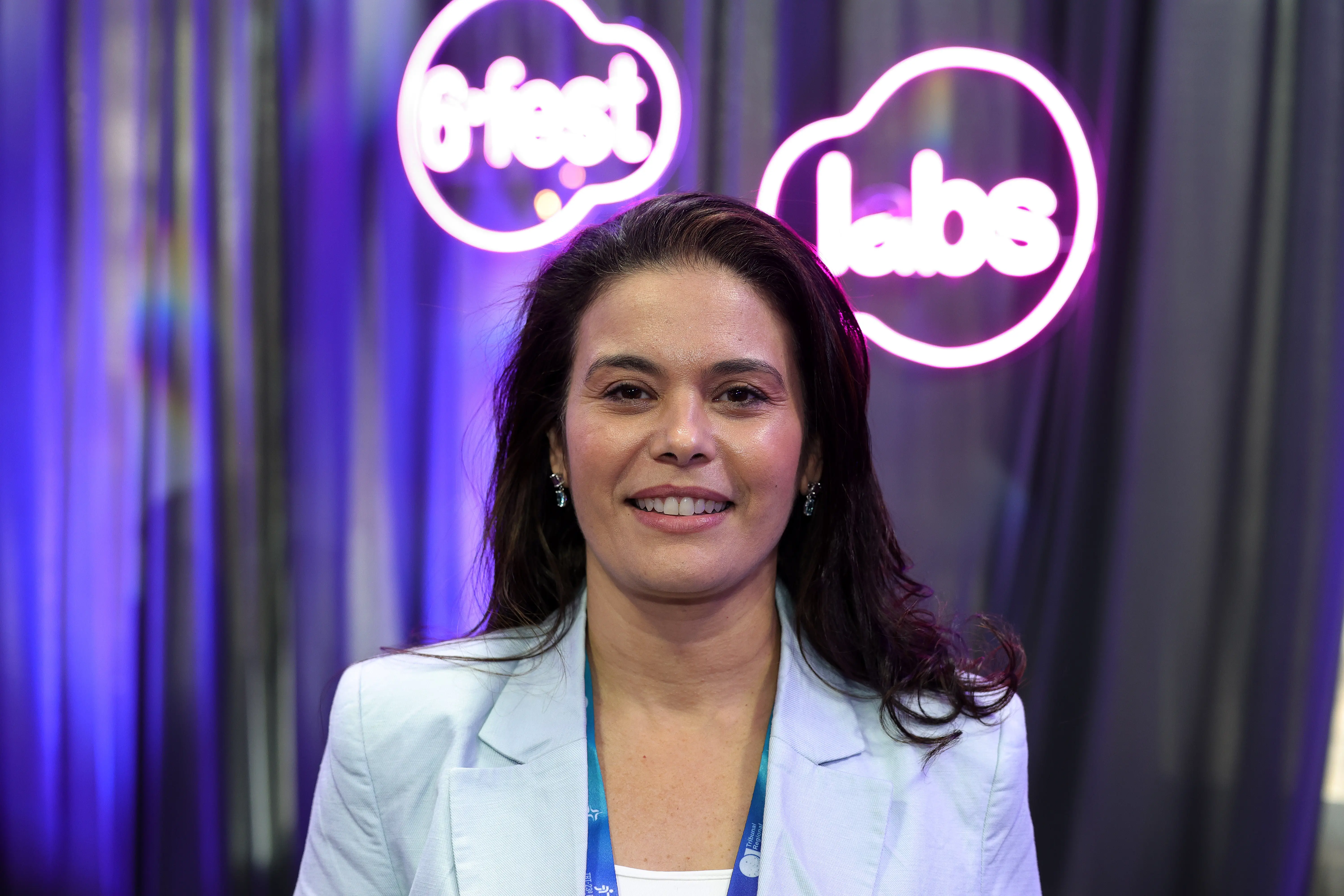 Retrato em plano médio de uma mulher de cabelos castanhos e blazer azul-claro sorrindo para a câmera. Ao fundo, uma cortina escura com neons luminosos nas cores rosa e roxo com a escrita "6º fest labs".