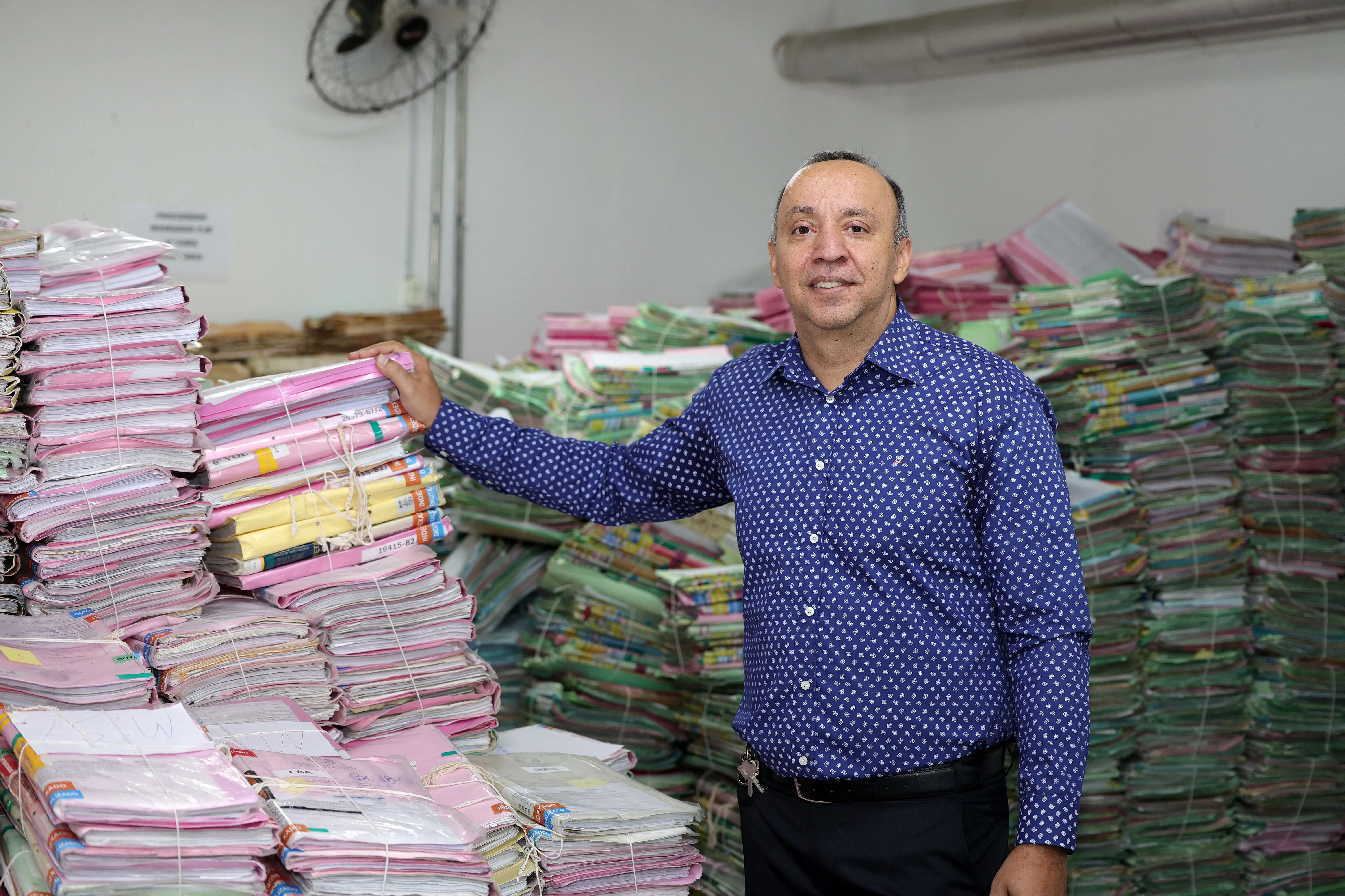 Um homem sorridente, de camisa azul com estampas brancas e calça escura, está em pé em um depósito, com a mão apoiada em uma das muitas pilhas de processos amarrados que lotam o ambiente ao seu redor. Ao fundo, há um ventilador de parede e incontáveis pilhas de documentos.