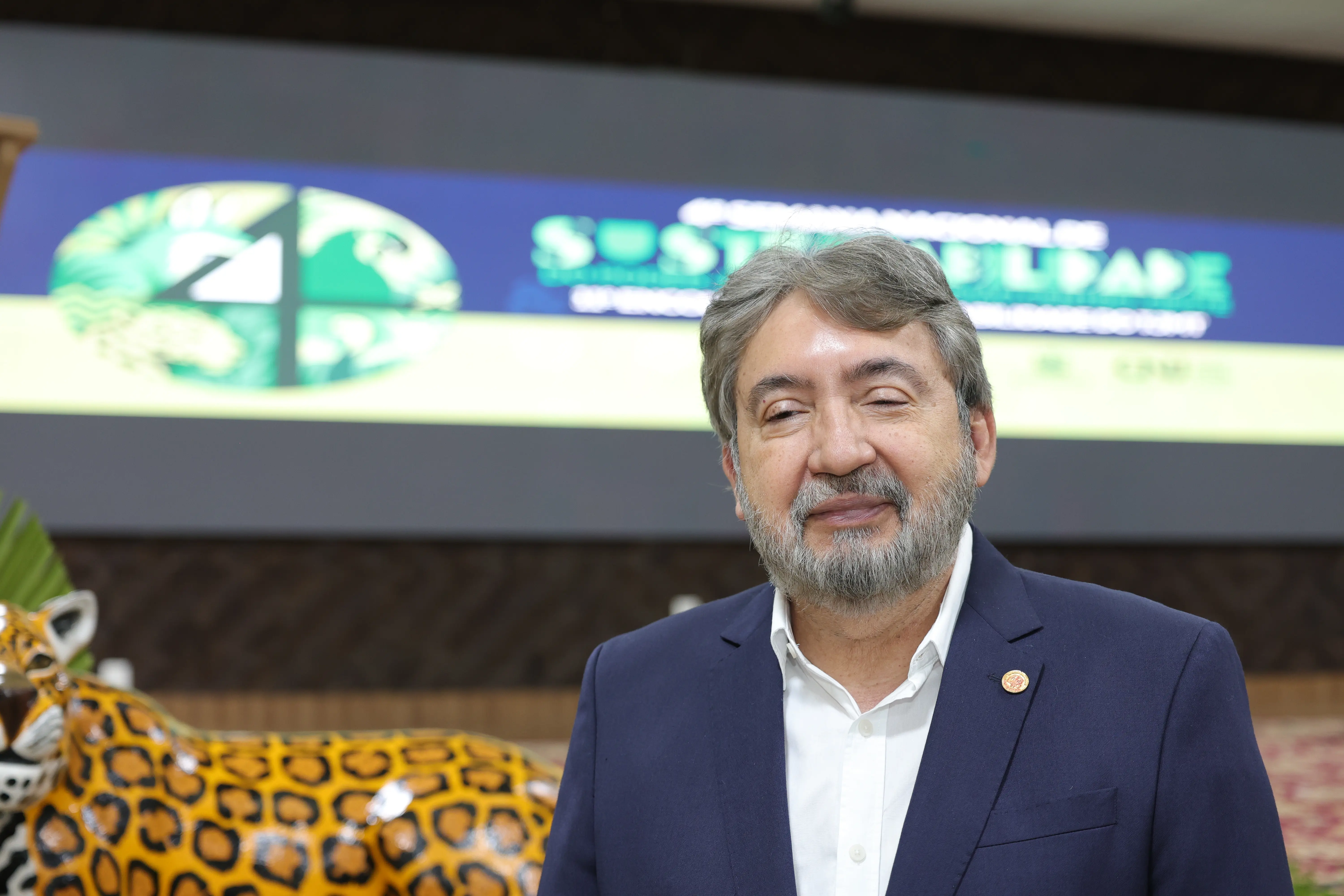 Um homem de barba e terno azul marinho posa na frente de um telão que exibe um logo verde e a palavra "SUSTENTABILIDADE". Uma estátua de onça-pintada está à esquerda.