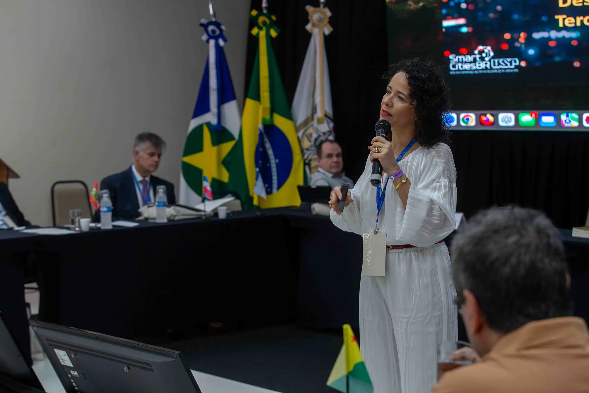 Foto que mostra a professora Ana Carla Bliacheriene proferindo palestra. ela está em pé, falando ao microfone. É uma mulher de pele clara, cabelos cacheados, escuros, na altura dos ombros, usando macacão branco. Em volta dela há uma mesa composta por diretores de escolas judiciais. 