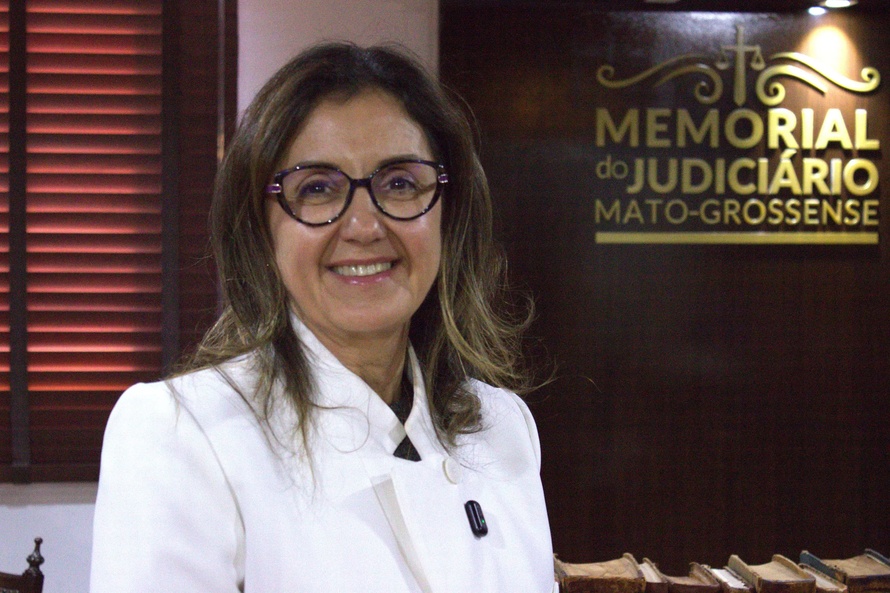 Mulher de cabelos castanhos ondulados, óculos e blazer branco sorri para a câmera. Ao fundo, painel iluminado do Memorial do Judiciário Mato-Grossense.