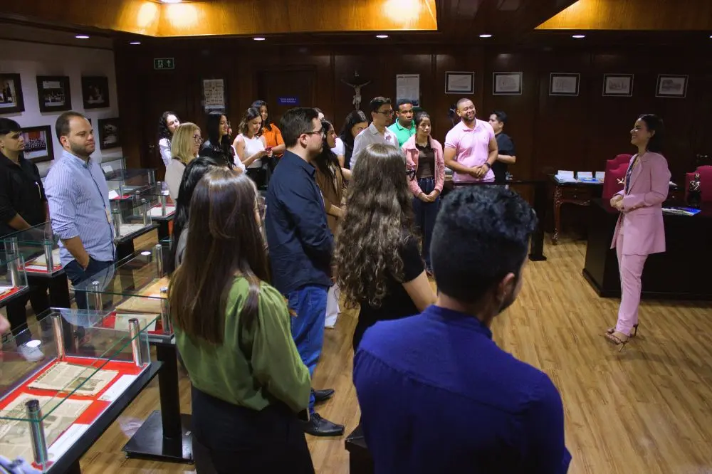 Foto horizontal em plano aberto que mostra um grupo de estudantes de direito observando uma mulher de calça e blazer cor-de-rosa, que está à frente falando. Eles estão no Espaço Memória do Tribunal de Justiça de Mato Grosso, local cheio de quadros e mesas de vidros com documentos antigos expostos.