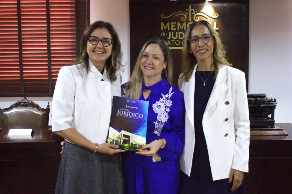 Foto horizontal que mostra três mulheres loiras posando para a foto. Elas estão sorrindo e duas delas seguram um exemplar do Glossário Jurídico do Tribunal de Justiça de Mato Grosso. Tratam-se de uma juíza e duas professoras de Direito. Elas estão no Espaço Memória do TJMT.