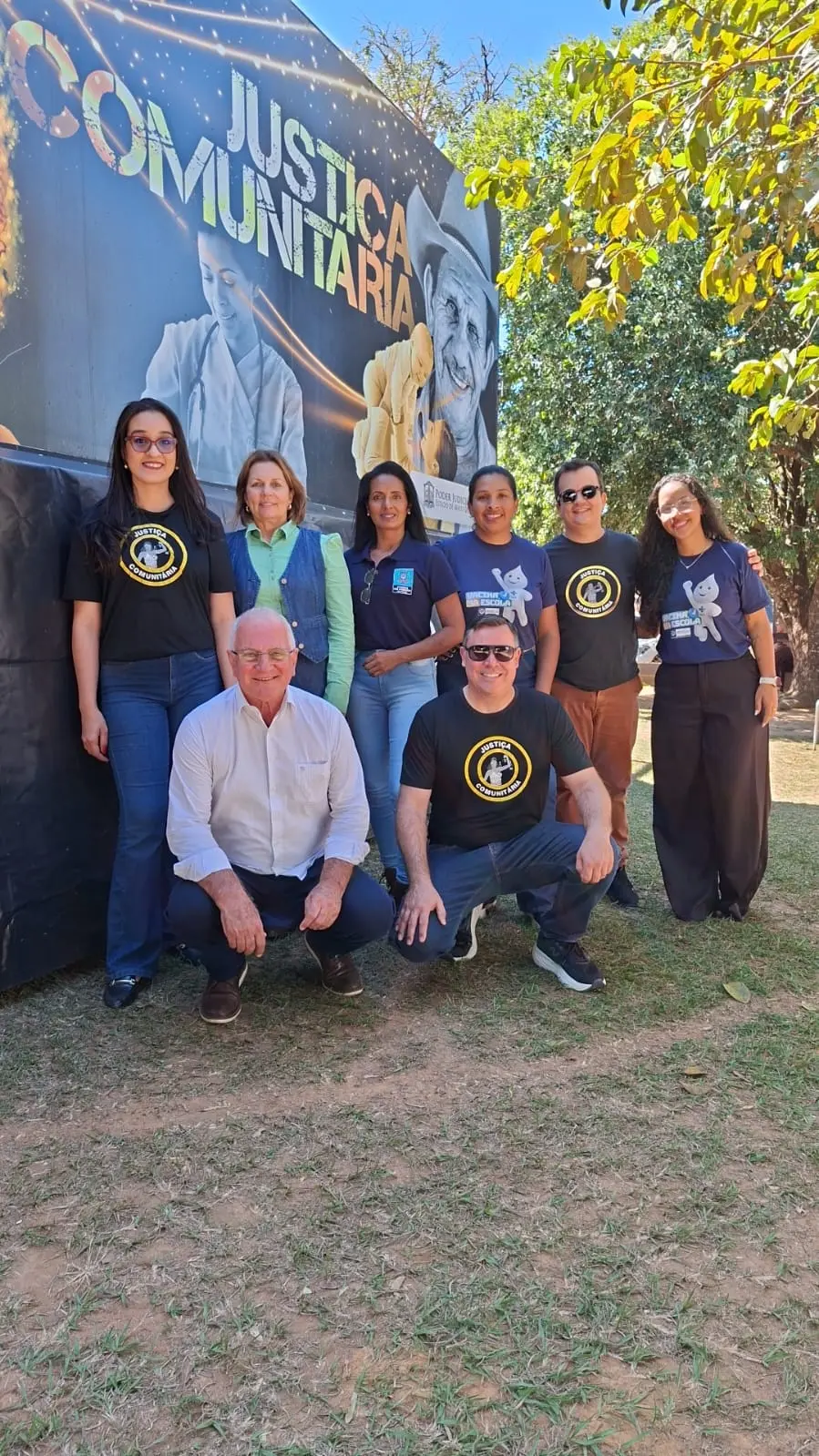 Foto vertical que mostra um grupo de seis mulheres e dois homens posando para a foto em frente à carreta da Justiça Comunitária, em um campo aberto com grama e árvores.