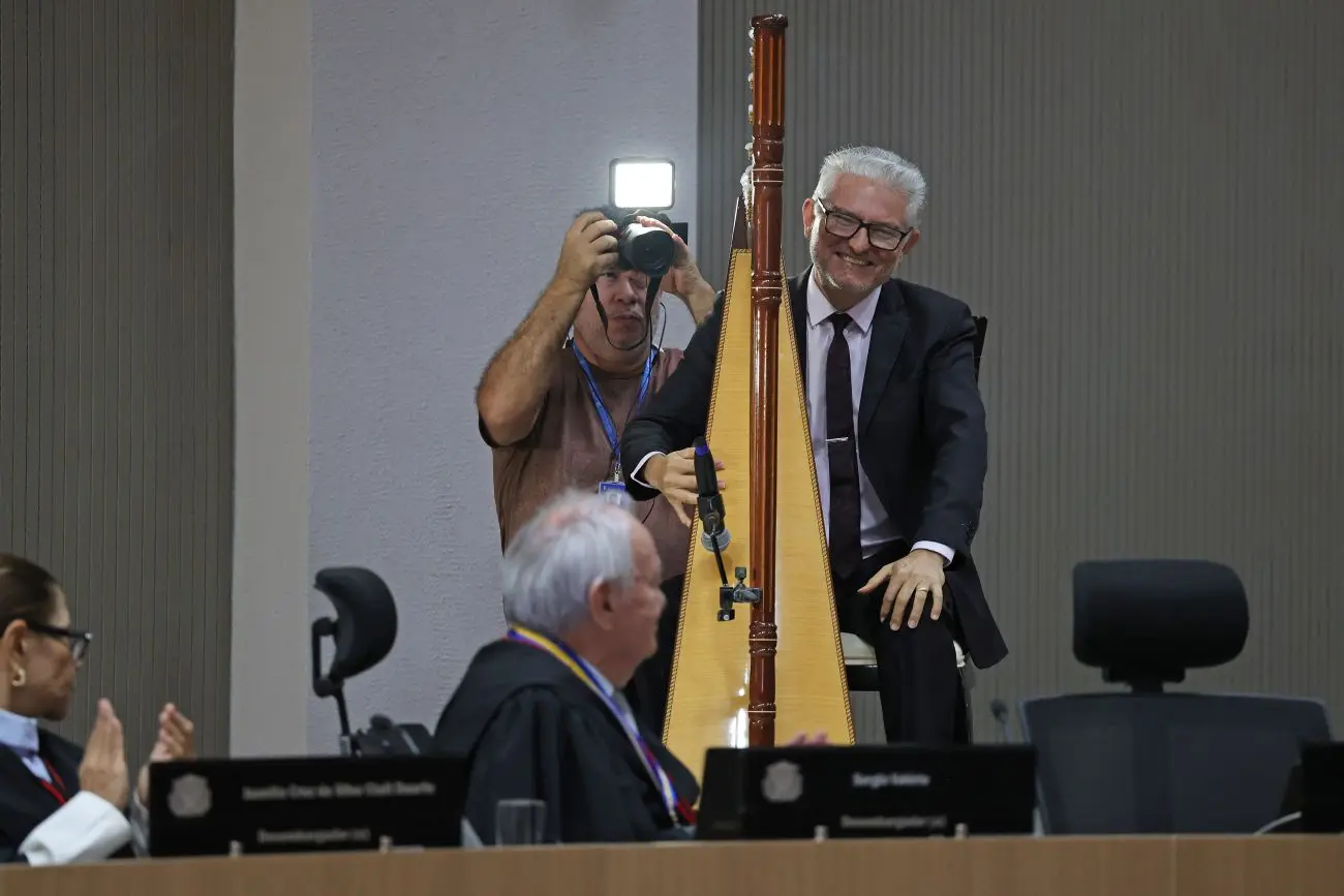 Foto horizontal que mostra o desembargador Sérgio Valério sentado em sua poltrona no plenário do TJMT, virado para trás, olhando para o harpista Narcizo Lucena, que está atrás dele, sorrindo em frente de uma harpa.