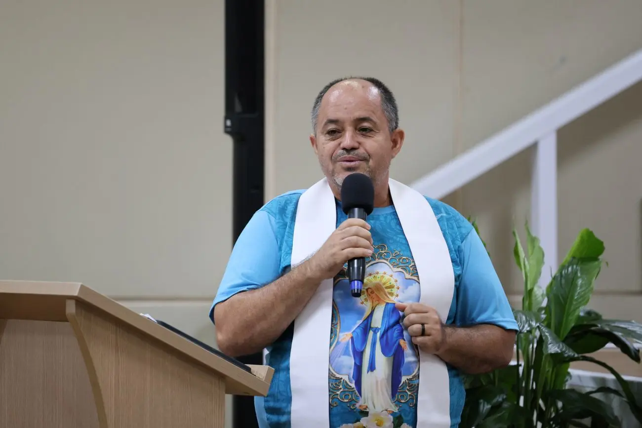 Foto horizontal que mostra o padre Divair Bento, da cintura pra cima, falando ao microfone, no auditório do TJMT, usando camiseta azul com estampa de Nossa Senhora e estola sacerdotal branca. Ele é um homem de baita estatura, calvo e de pele parda.