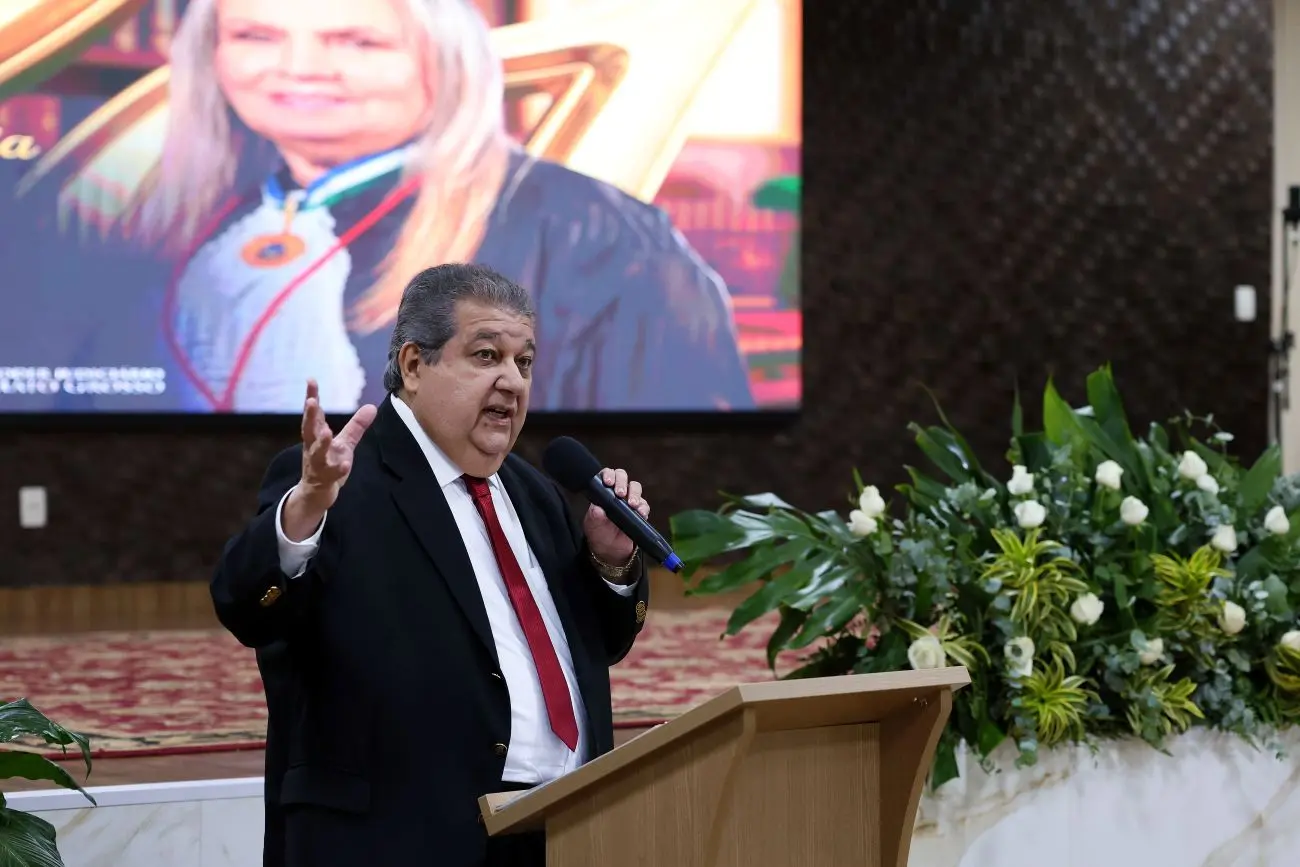 Foto horizontal que mostra o procurador de justiça Paulo Prado falando ao microfone e gesticulando, em pé, no auditório do TJMT. Ele é um homem branco, obeso, de cabelos grisalhos, usando camisa branca, gravata vermelha e terno preto. Ao fundo, o telão exibe foto da desembargadora Maria Erotides.