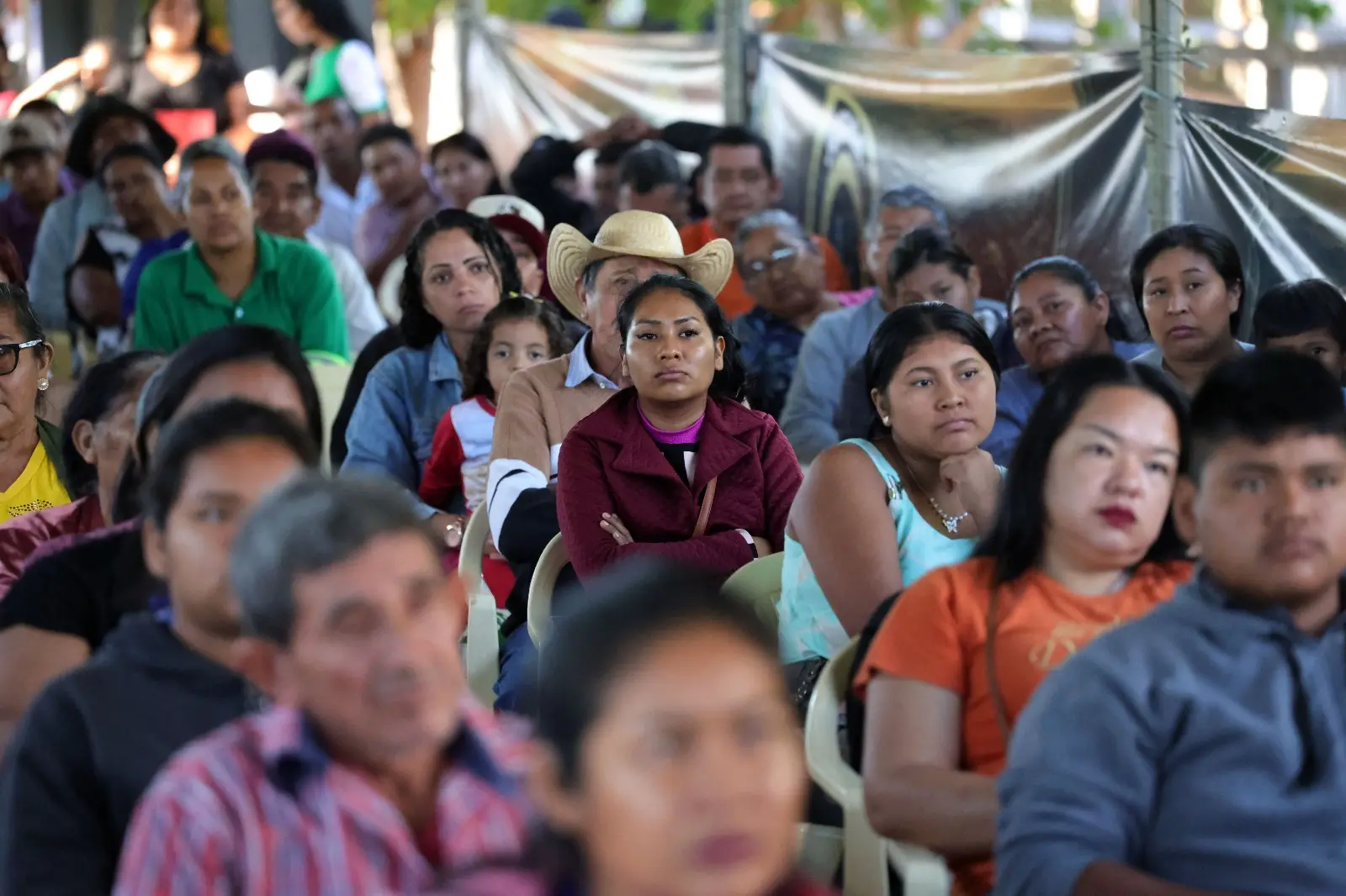 Público diverso, majoritariamente indígena, assiste a um evento. Sentadas em cadeiras plásticas, as pessoas parecem atentas. Em destaque no centro, uma mulher de jaqueta grená e braços cruzados.