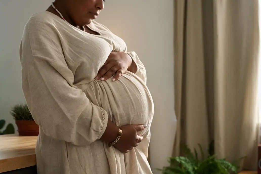 Mulher negra vestindo vestido bege de mangas compridas segura a barriga de grávida com as duas mãos. O enquadramento vai do peito ao quadril. Ela usa colar e pulseira. Ao fundo, há uma janela com cortina clara, um móvel de madeira e plantas em vasos.