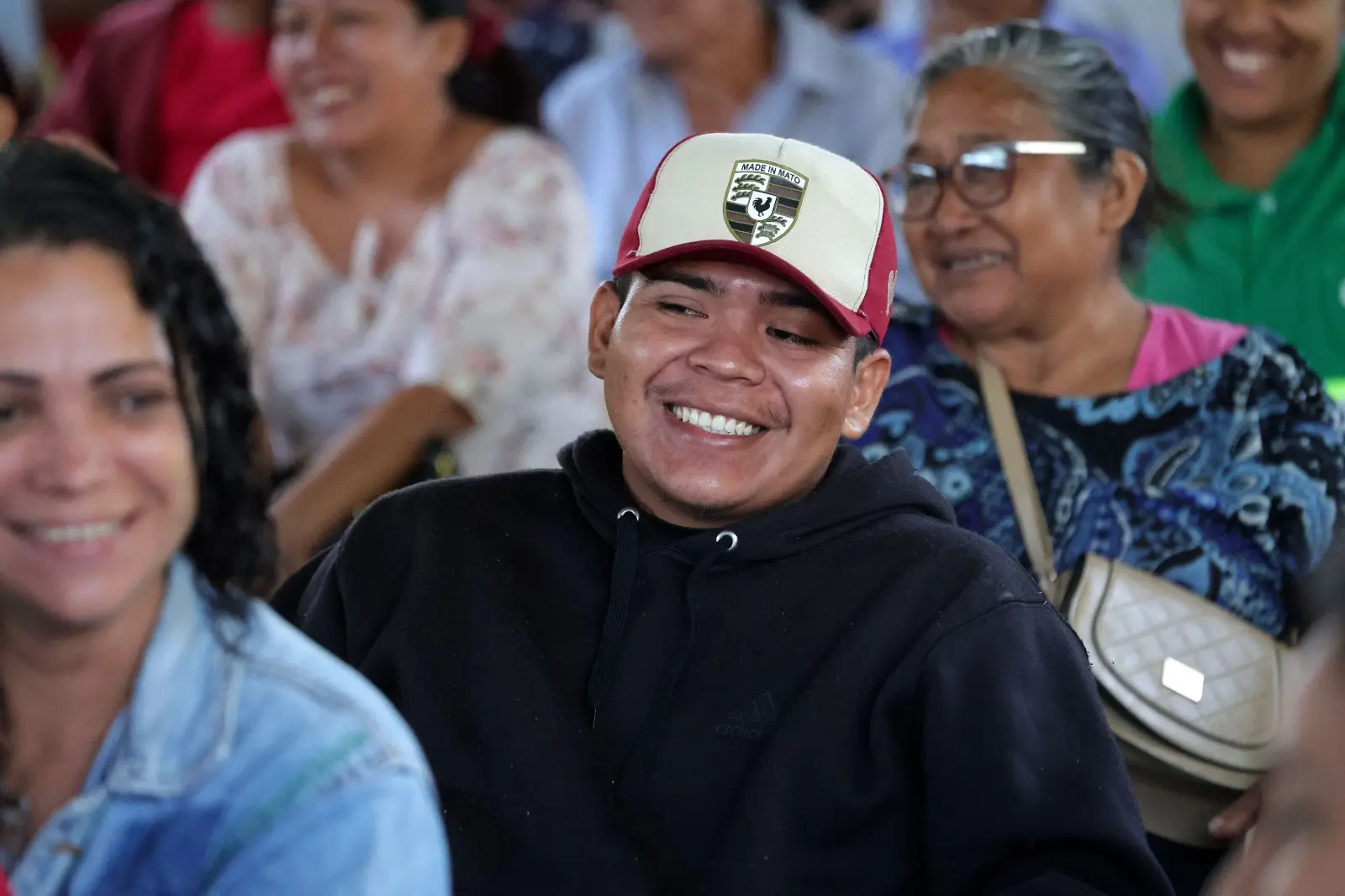 Jovem sorridente e com dentes à mostra, vestindo capuz preto e boné bege com aba e detalhes em bordô, é o foco da imagem. Ele está cercado por outras pessoas felizes, desfocadas em segundo plano.