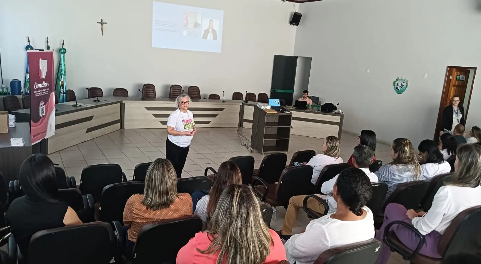 Mulher de cabelos grisalhos e óculos, veste camiseta branca e calça preta, fala em pé no auditório. Ao fundo, plateia sentada assiste à palestra sob banner da Cemulher. 