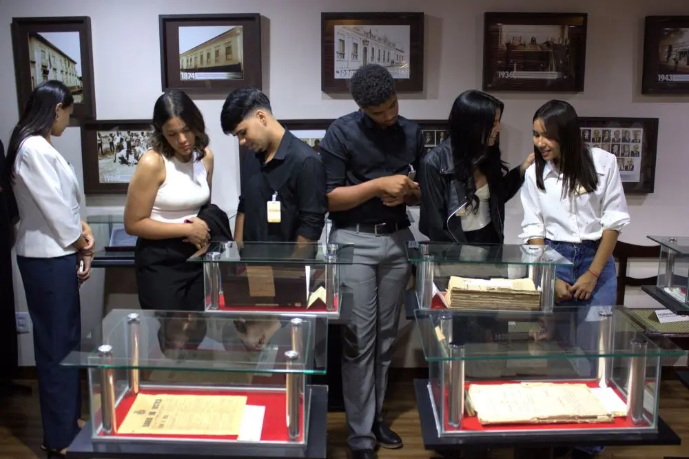 Foto horizontal em plano aberto que seis estudantes de Direito (sendo dois rapazes ao centro e quatro moças) observando documentos antigos, expostos em mesas de vidro, no Espaço Memória do Tribunal de Justiça de Mato Grosso. Atrás deles, uma parede com vários quadros com fotos antigas do Tribunal.