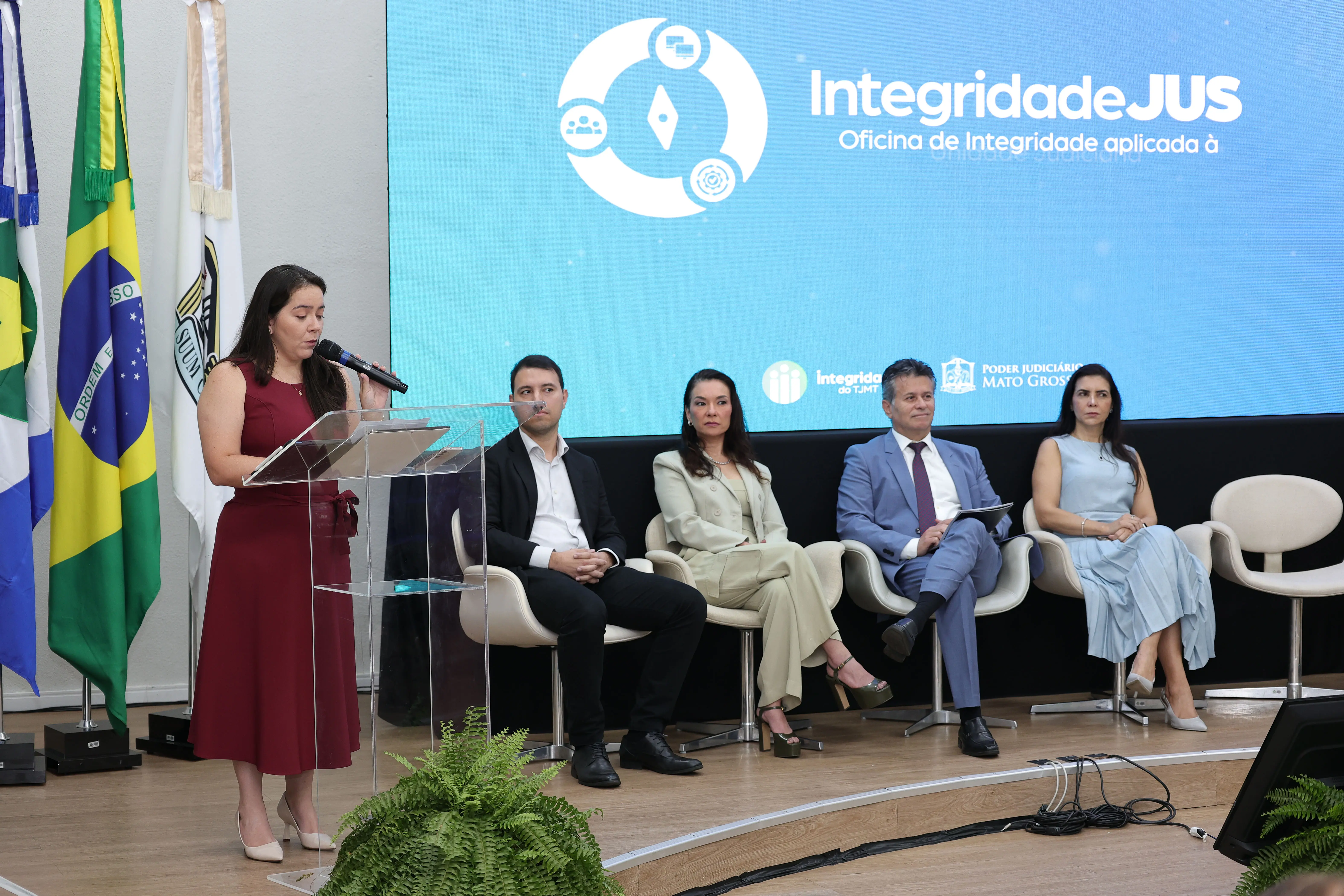 Mulher de vestido vinho fala ao microfone em um púlpito transparente. Ao lado, autoridades sentadas em poltronas observam no palco diante de telão azul com a marca "IntegridadeJUS".