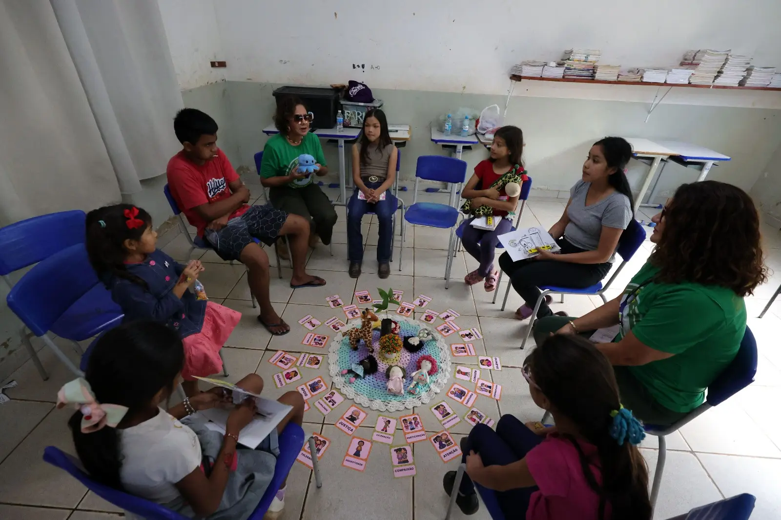 Uma roda de conversa em uma sala de aula simples, onde crianças e duas mulheres adultas estão sentadas em cadeiras azuis. No centro, sobre um tapete circular, há brinquedos e cartões de atividades.
