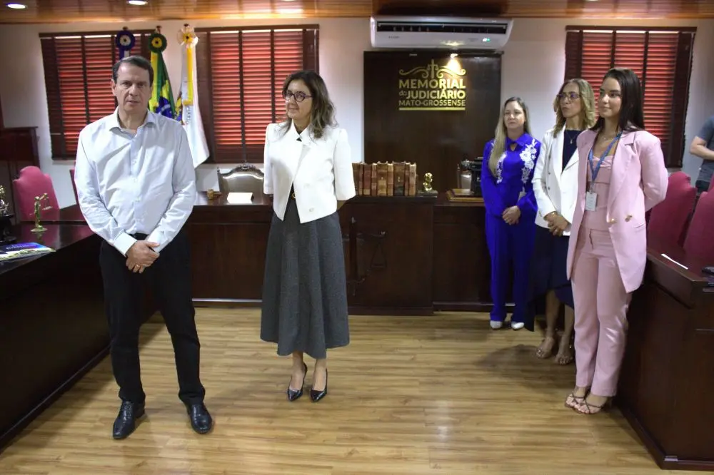 Foto horizontal em plano aberto que mostra um servidor e uma juíza do TJMT em pé ao centro do Espaço Memória do Tribunal. No lado direito da foto, três mulheres estão em pé. Todos usam roupa formal e o ambiente é clássico, com móveis de jacarandá antigos, cadeiras com estofado vermelho e persianas.