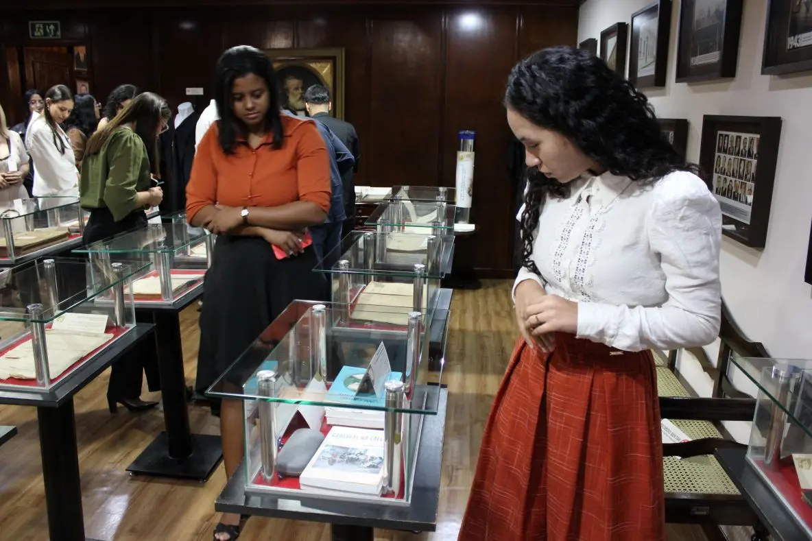 Foto horizontal em plano aberto que mostra estudantes de Direito observando documentos antigos, expostos em mesas de vidro, no Espaço Memória do Tribunal de Justiça de Mato Grosso. Atrás deles, uma parede com vários quadros com fotos antigas do Tribunal.