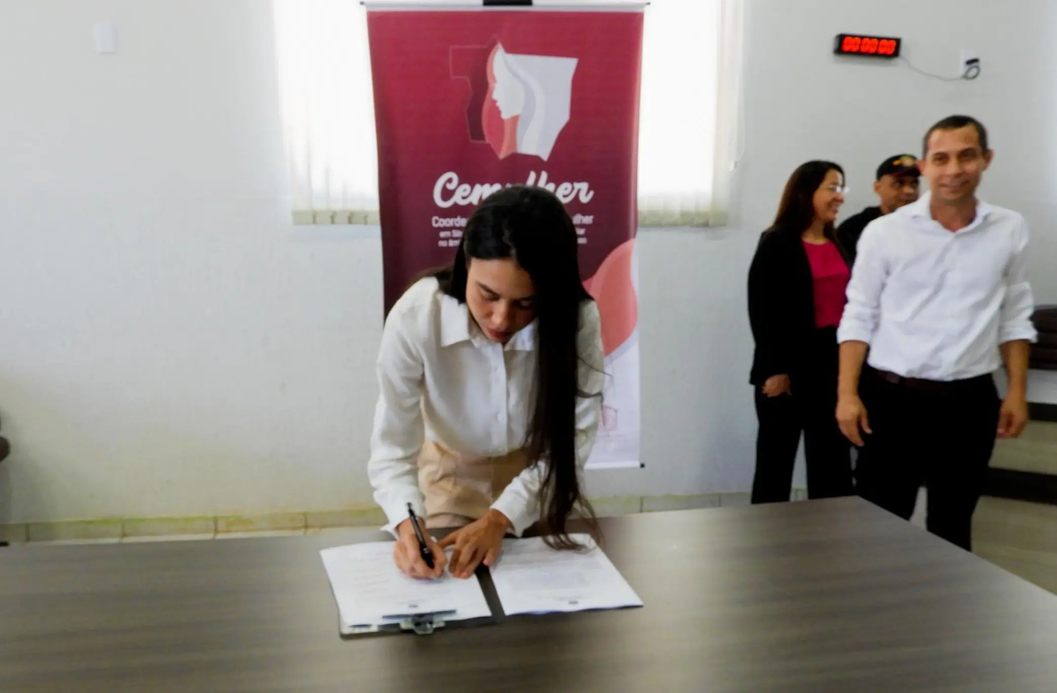 Mulher de cabelos longos e escuros, veste blusa branca e saia bege, assina documento sobre a mesa. Ao fundo, banner da Cemulher e dois homens observam a cena. 