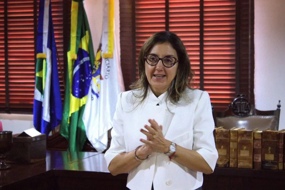 Foto horizontal da juíza Christiane da Costa Marques falando com semblante alegre e as mãos juntas à frente do corpo. Ela é uma mulher branca, loira com cabelo na altura dos ombros, usando camisa branca e óculos de grau. O ambiente é um gabinete com três mastros com bandeiras e livros antigos.