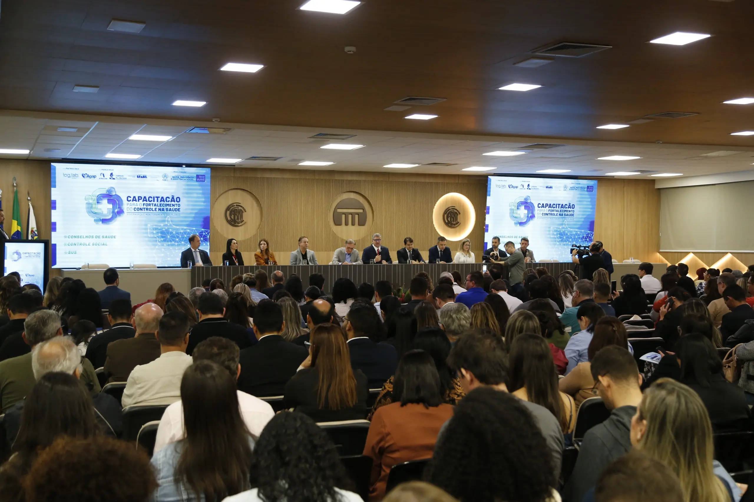 Foto horizontal em plano aberto que mostra o auditório do Tribunal de Contas lotado de pessoas sentadas. Á frente, no palco, diversas autoridades sentadas.
