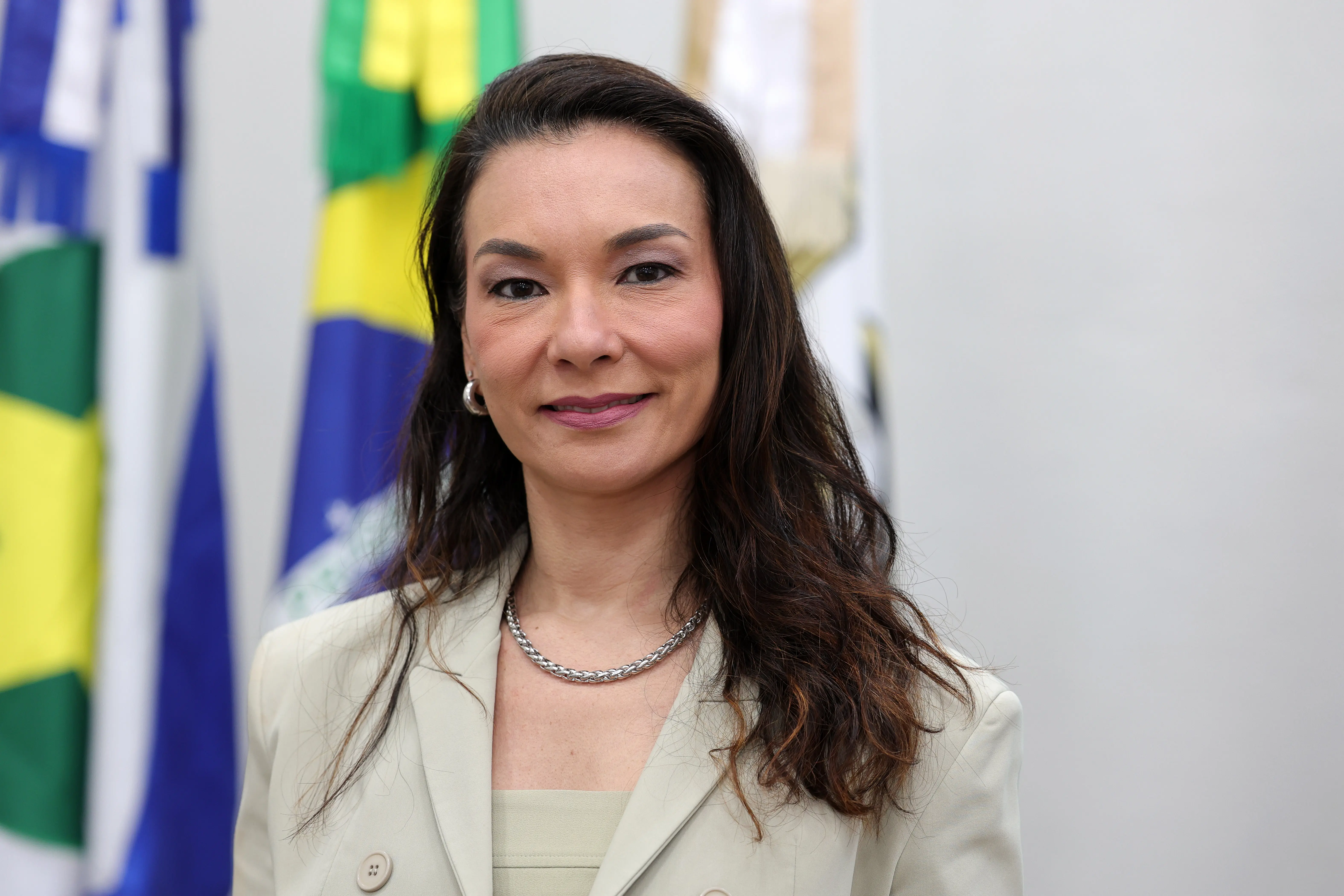 Retrato da juíza Hanae Yamamura, que é uma mulher com traços orientais, pele clara e longos cabelos castanhos, veste um blazer bege sobre um top da mesma cor e um colar de corrente. Ela sorri suavemente para a câmera, com bandeiras desfocadas ao fundo.