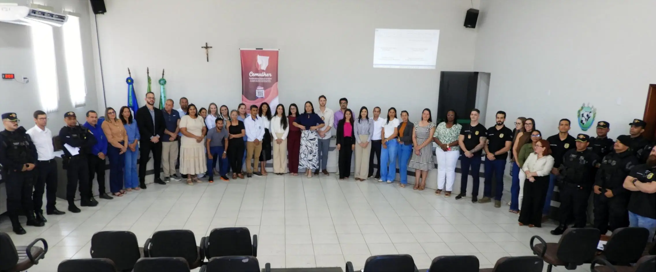 Grande grupo de pessoas, entre servidores e policiais, posa enfileirado no auditório. Ao fundo, banner da Cemulher, bandeiras e um crucifixo na parede.