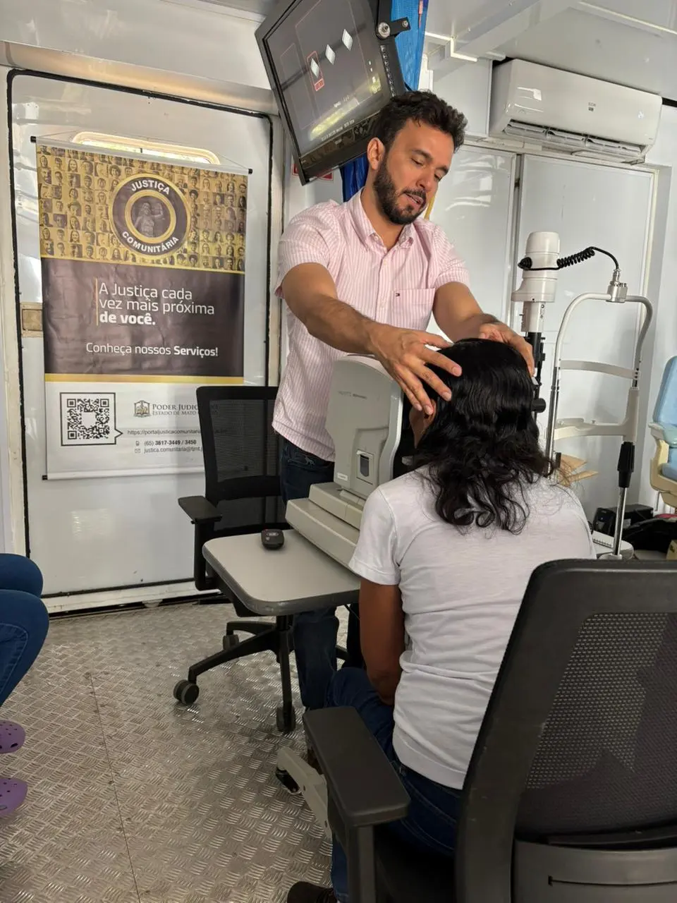 Foto vertical que mostra um homem ajeitando a cabeça de uma mulher no aparelho para exame de vista, dentro da carreta de atendimento oftalmológico da Justiça Comunitária.