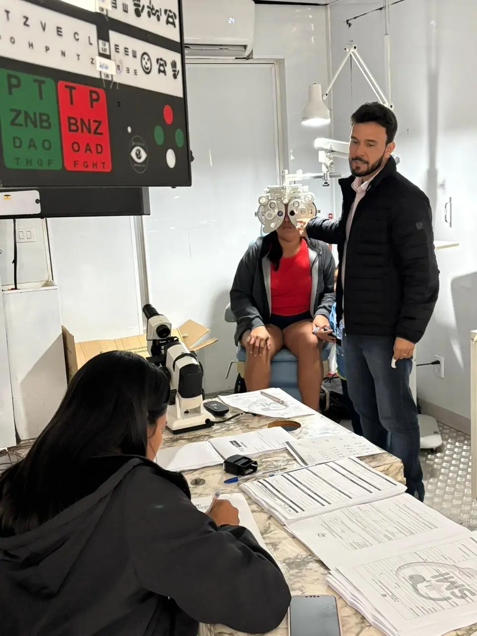 Foto vertical que mostra um homem atendendo uma mulher no aparelho para exame de vista, dentro da carreta de atendimento oftalmológico da Justiça Comunitária. Outra mulher, sentada, faz anotações. 