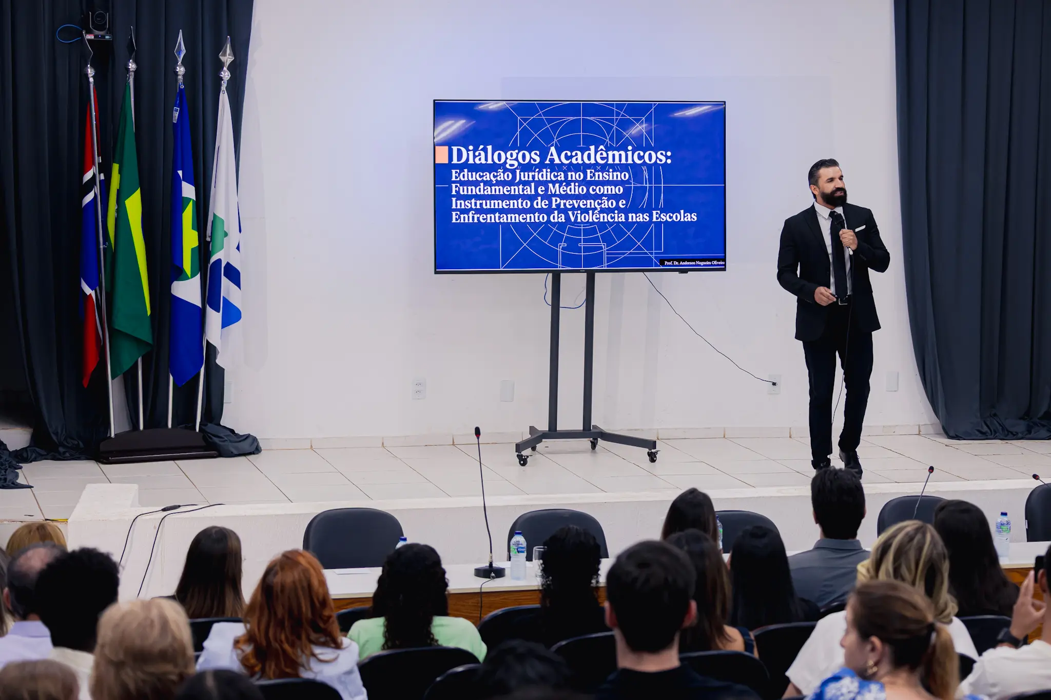 Homem de terno preto e barba fala ao microfone em palco ao lado de TV com slide azul sobre educação jurídica. À esquerda, quatro bandeiras oficiais; à frente, a plateia de costas.